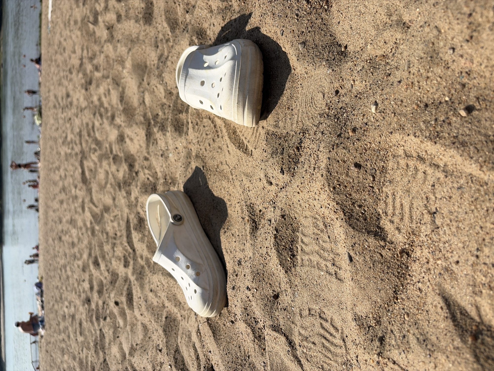 The image depicts a sandy beach with two white Crocs shoes placed on the sand in the foreground. In the background, a body of water, possibly a river, is visible with several people scattered along the shoreline, some of whom are in the water or sitting on the sand. The scene suggests a recreational area, typical of a summer day. The location is identified as Omsk, Russia.