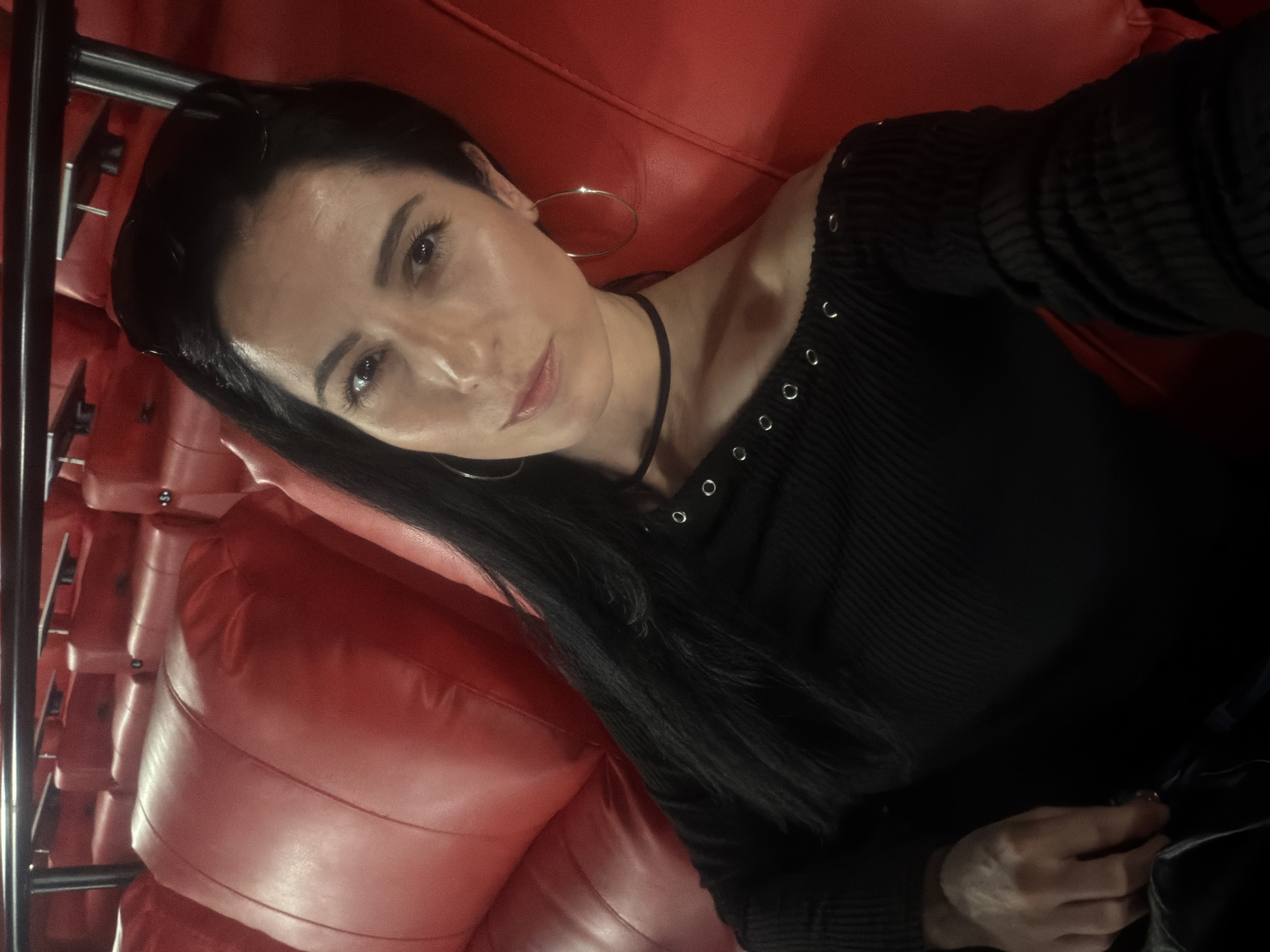 A woman with dark hair is lying on a red leather couch. She is wearing a black off-the-shoulder top and hoop earrings. The couch appears to be part of a seating arrangement with metal framing. The location is potentially in Pleven, Bulgaria.