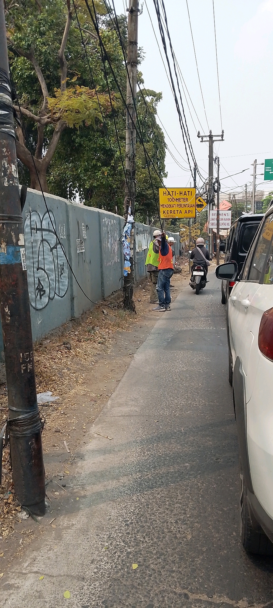 The image depicts a street scene in Bekasi, Indonesia, near a railway crossing. In the foreground on the left is a weathered, dark metal pole with some black tape wrapped around its base. To the right of the pole is a paved sidewalk with dry leaves scattered on it. Behind the sidewalk is a tall, gray concrete wall covered in graffiti. Several power lines crisscross above the scene, attached to wooden utility poles.
In the mid-ground, three individuals are present. Two are wearing high-visibility vests, one yellow and one orange, along with hard hats and jeans, suggesting they are construction or road workers. The person in the orange vest is standing near a utility pole, holding something in their hand and looking upwards. The third person is wearing a helmet and a gray jacket, riding a motorcycle that is heading away from the camera and towards the right side of the image.
A yellow rectangular sign with black text is prominently displayed on a utility pole. The sign reads "HATI-HATI 100 METER MENDEKATI PERINTAS KERETA API", which translates to "CAUTION 100 METERS APPROACHING RAILWAY CROSSING". Further down the road, other signs are visible, including one that appears to be a stop sign.
The background consists of trees with lush green foliage and a clear sky. To the right of the motorcycle, a white car is partially visible, along with the rear of a dark-colored vehicle in front of it. The overall scene suggests a working area near a transportation route.
