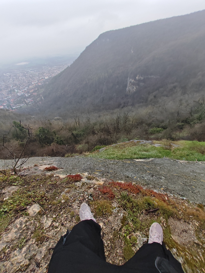 The image displays a first-person perspective from an elevated position, likely the Shumen Plateau overlooking the city of Shumen, Bulgaria. In the immediate foreground, the lower legs and feet of an individual are visible, clad in black pants and light pink athletic shoes. The feet are positioned near a rocky edge, which is covered with sparse green vegetation, moss, and distinctive patches of red, succulent-like ground cover. Bare tree branches are visible on the left edge of the foreground. Beyond this immediate terrain, a steep, densely forested mountainside descends into the valley. The trees are predominantly bare deciduous, indicating late autumn or early spring conditions. Further in the background, the city of Shumen spreads across the valley floor, characterized by numerous residential and commercial structures with varying rooftop colors, appearing somewhat indistinct due to distance and atmospheric haze. A lighter, flat expanse is visible on the horizon beyond the city. The sky is uniformly overcast and grey, contributing to muted lighting across the scene. No other individuals or explicit actions beyond observation are depicted.