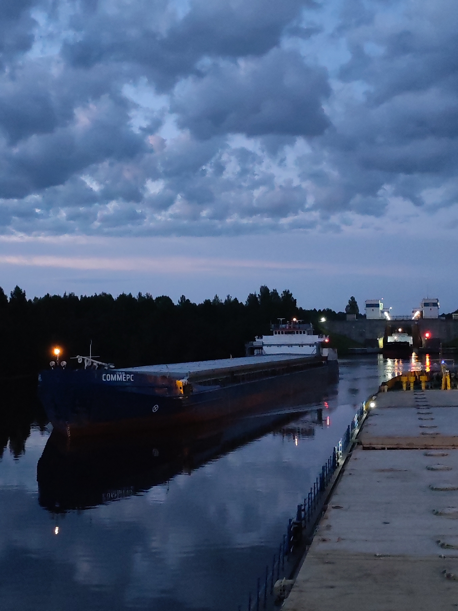The image depicts a dusk scene at the Devyatiny, Russia water lock. A large barge, identified by the name "COMMERC" on its hull, is positioned in the canal. The barge is dark blue and appears to be loaded. To the right of the barge, a concrete dock with metal bollards is visible. Further in the background, structures resembling lock gates are present, with lights illuminating the water. A second, smaller white vessel is also visible within the lock system. The sky is overcast with dark, dramatic clouds, and the water reflects the dim light and the surrounding environment. A line of dark trees forms the background on the left side of the image.