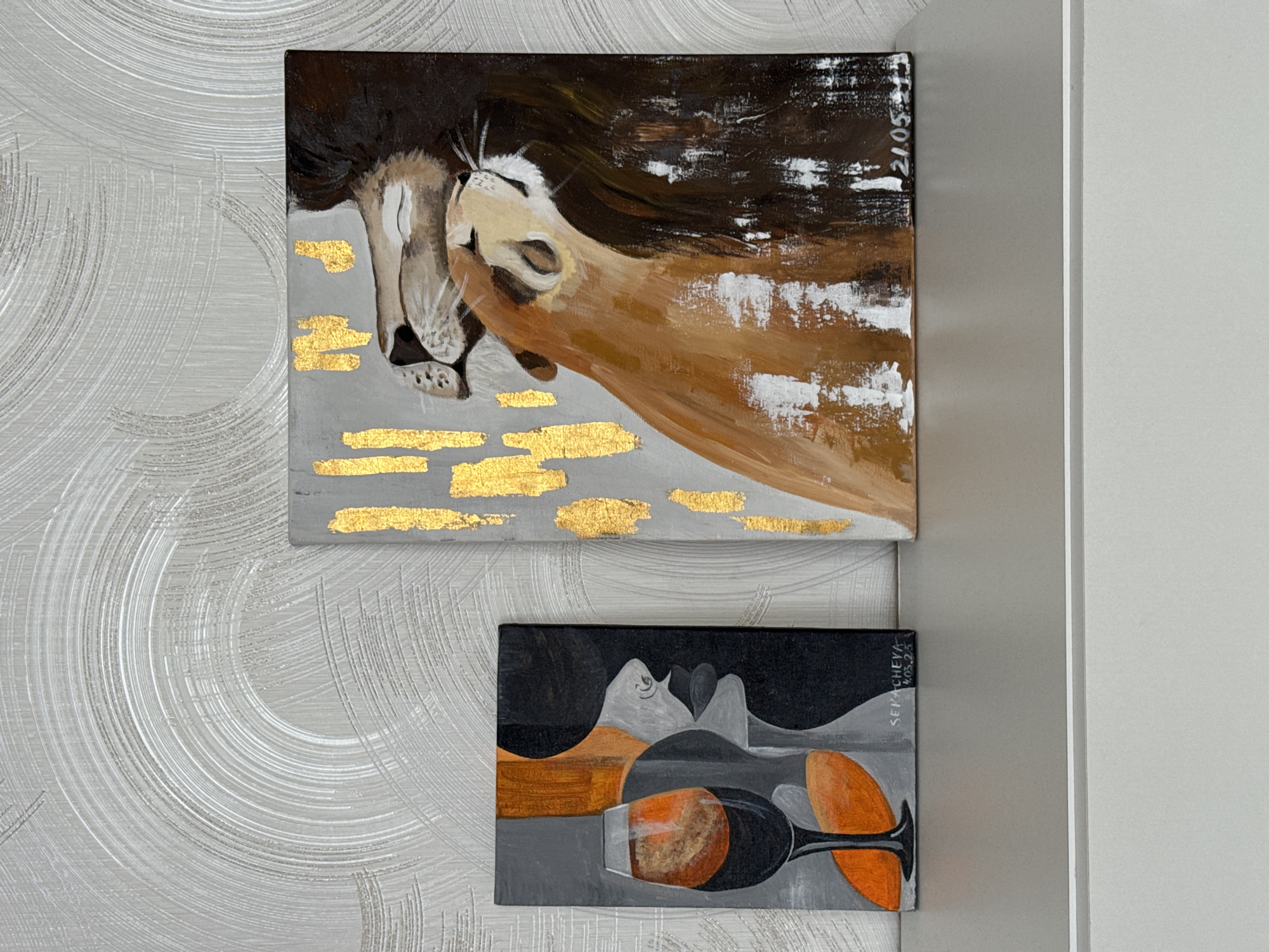 Two paintings hang on a textured light grey wall next to a white door frame. The upper painting depicts a lion's head resting on its arm, rendered in brown, grey, white, and gold paint. The bottom painting is of a more abstract nature, displaying a black, grey, brown, and orange composition of what appear to be abstract people and glasses with a signature in the upper right corner. The paintings appear to be painted on canvases. There are no people visible. The location is unknown.