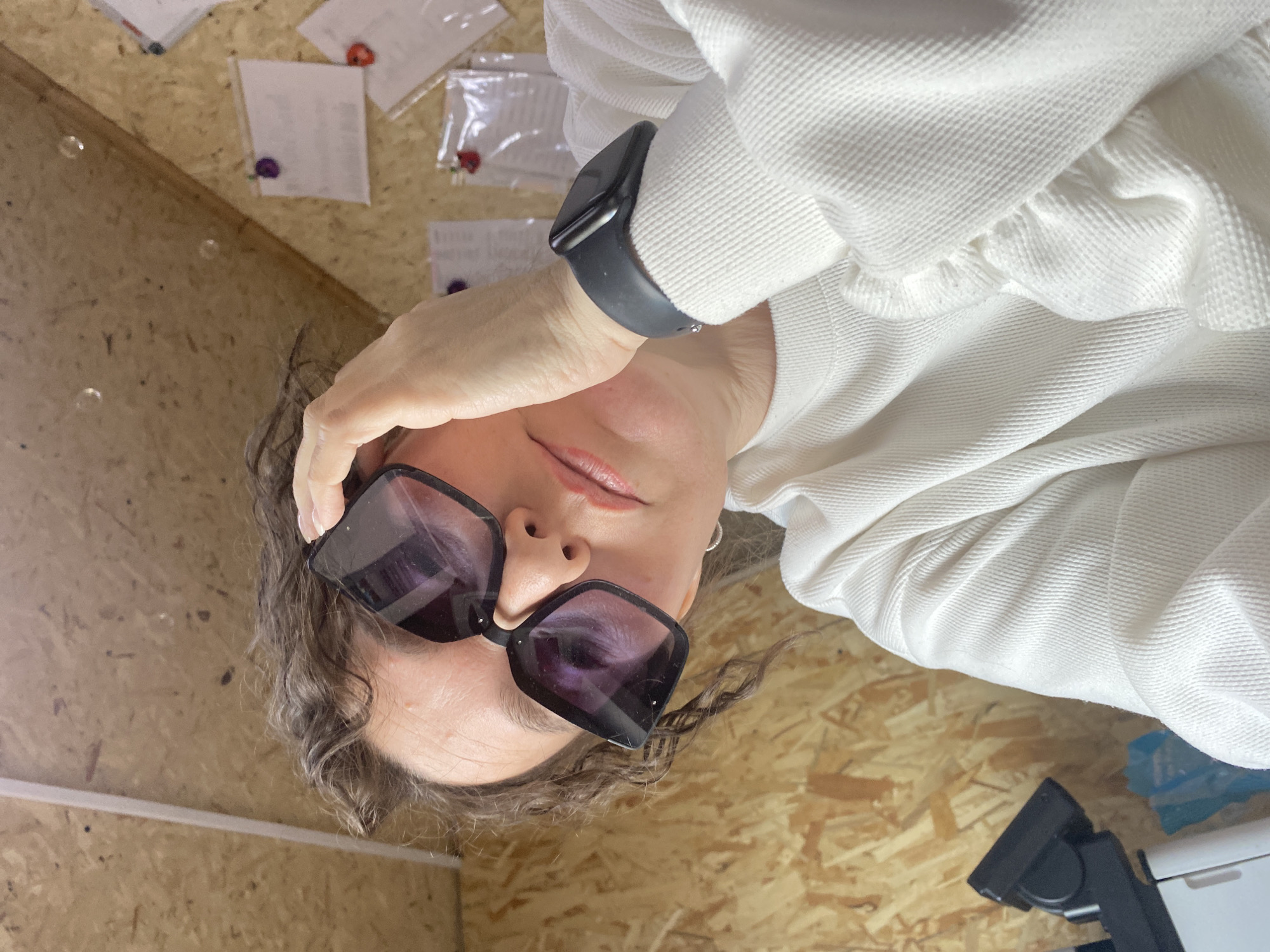 The image shows a woman situated indoors, specifically in what appears to be a room or attic with sloped, light-colored wooden wall paneling, identified as being in Ol'khovskoye, Russia. The woman, with curly brown hair, is seen from the chest up, gazing directly at the viewer. She wears large, black-framed sunglasses, with her right hand positioned at her temple, appearing to hold or adjust them. A dark smartwatch is visible on her left wrist. She is dressed in a white, long-sleeved sweater. In the background, several white paper notes, some enclosed in clear plastic, are affixed to the wooden wall using colorful thumbtacks (purple and red). A white vertical strip is present on the wall to the left. A portion of a black and white device, possibly a microscope, is visible on the lower left.