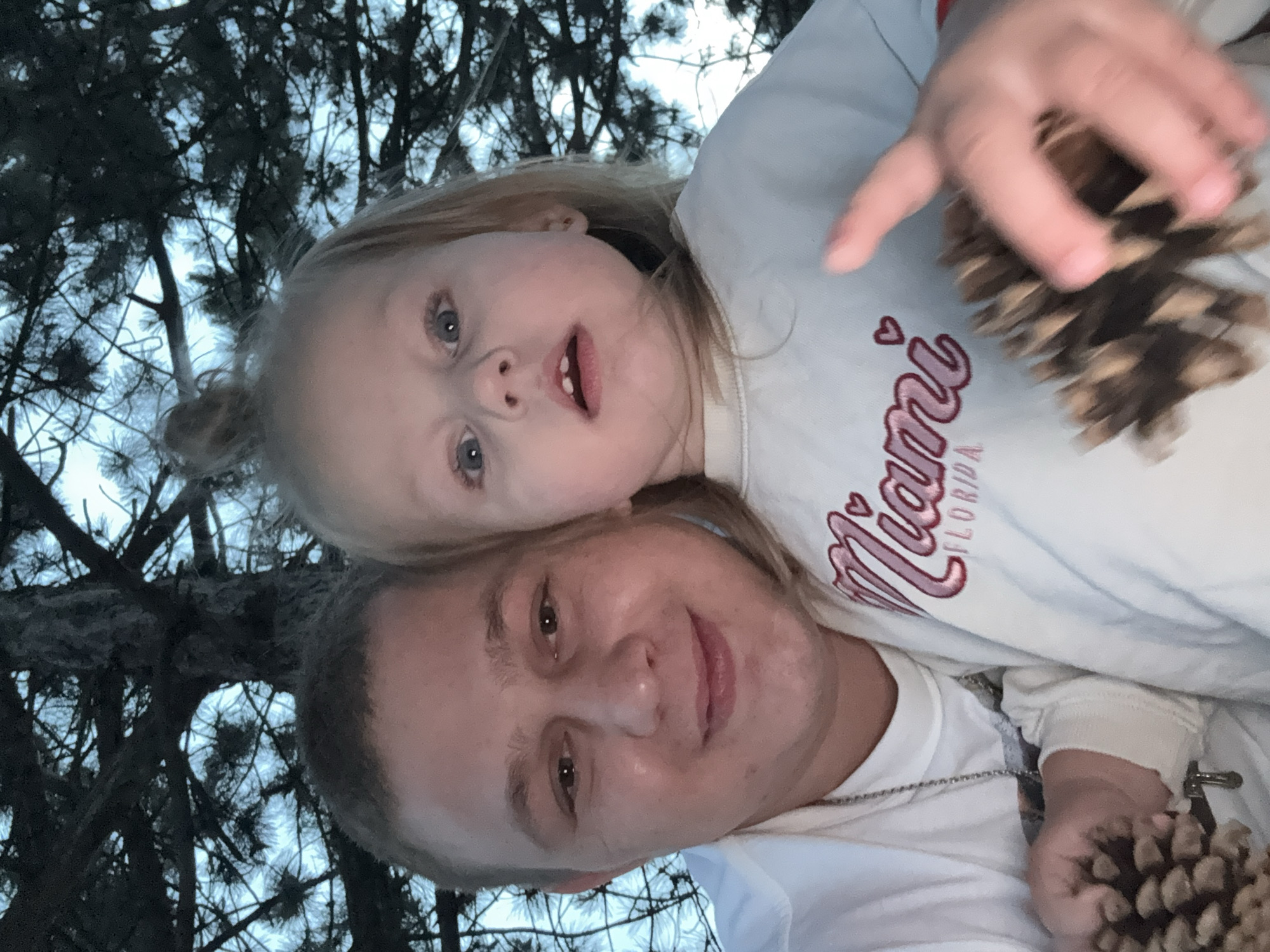 In Pilsen, Czechia, an outdoor selfie shows a fair-skinned male and a young fair-skinned female in close proximity, positioned in front of a dark evergreen tree with visible branches and needles against a twilight sky. The male, on the left, has short brown hair, a round face, and is smiling. He is wearing a white shirt and a silver chain necklace. The female, to the right, has blonde hair pulled into a bun and is looking directly at the camera with wide blue eyes. She is wearing a light-colored sweatshirt with the word "Miami" and "Florida" embroidered in pink. Both individuals are holding pinecones. The male holds a pinecone in his left hand, and the female's right hand is near a pinecone that is partially obscured by her movement, suggesting she may be interacting with it. The overall lighting indicates it is dusk or a shaded area.