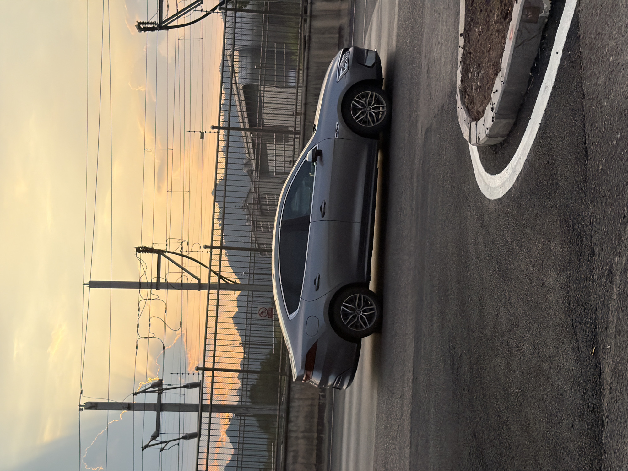 A gray sedan drives on a road with mountains and overhead power lines in the background during sunset. The car is positioned in the center of the frame, moving from left to right. The sky is filled with a soft orange and yellow glow from the setting sun, with rays of light visible through wispy clouds. A metal fence runs horizontally across the middle ground, separating the road from the area behind it, which includes the mountains and some structures. The foreground shows a portion of the asphalt road and a newly constructed curb with soil and rocks. This scene is located in the city of Jiangjin, China.