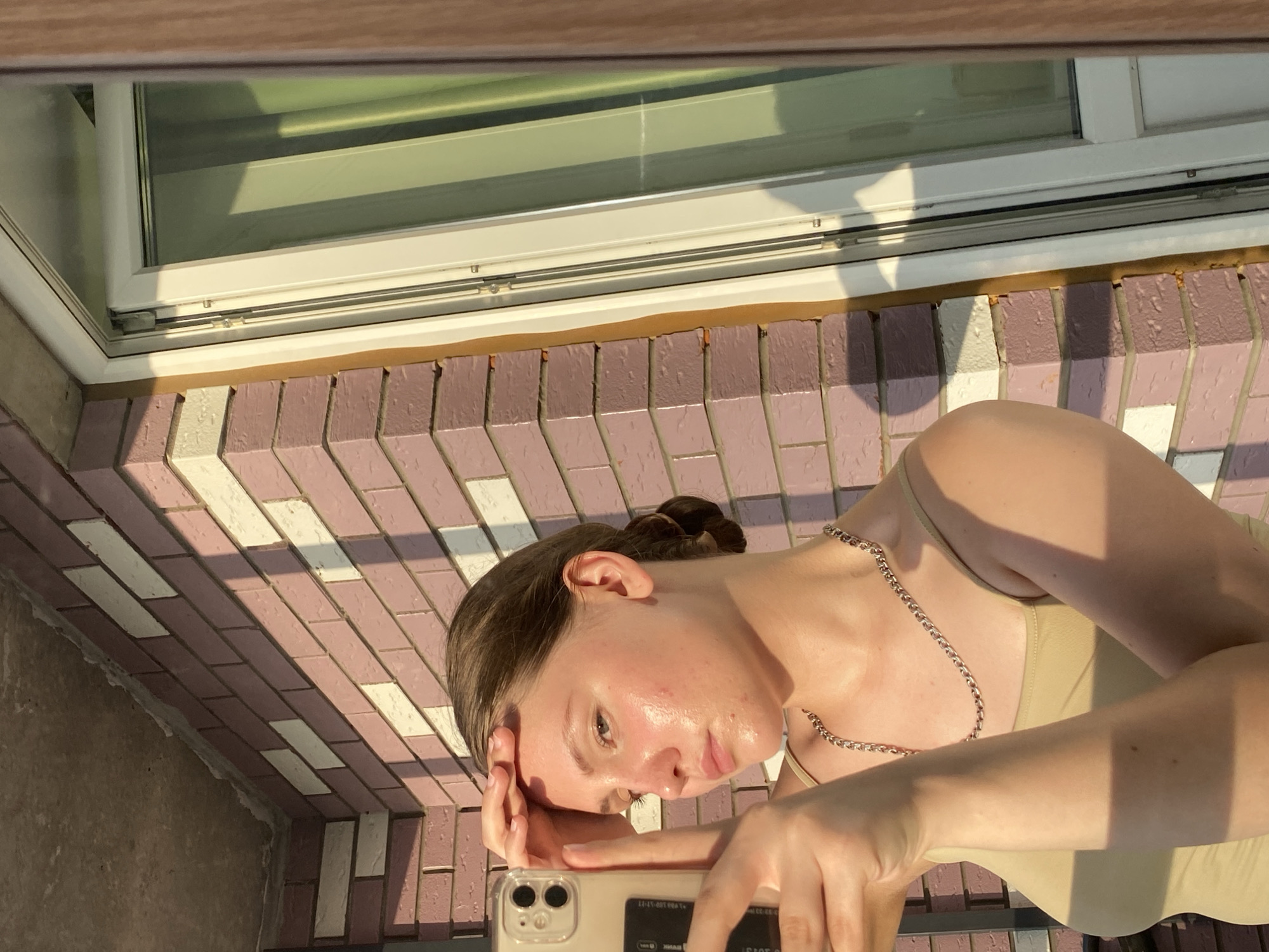 An outdoor, waist-up selfie shows a young woman with light brown hair pulled into a bun, wearing a beige tank top and a silver chain necklace. She is holding her left hand to her forehead, with her phone visible in her right hand, angled to capture her reflection. Her skin appears dewy, reflecting the warm, golden light of what appears to be late afternoon or early evening sun. Behind her, a brick wall with alternating rows of pink and white bricks creates a patterned backdrop. To her right, a white-framed window with reflective glass is partially visible. The scene suggests a location in Izhevsk, Russia, possibly on a balcony or near an exterior wall of a building. The overall lighting and shadows indicate an outdoor setting with strong sunlight.