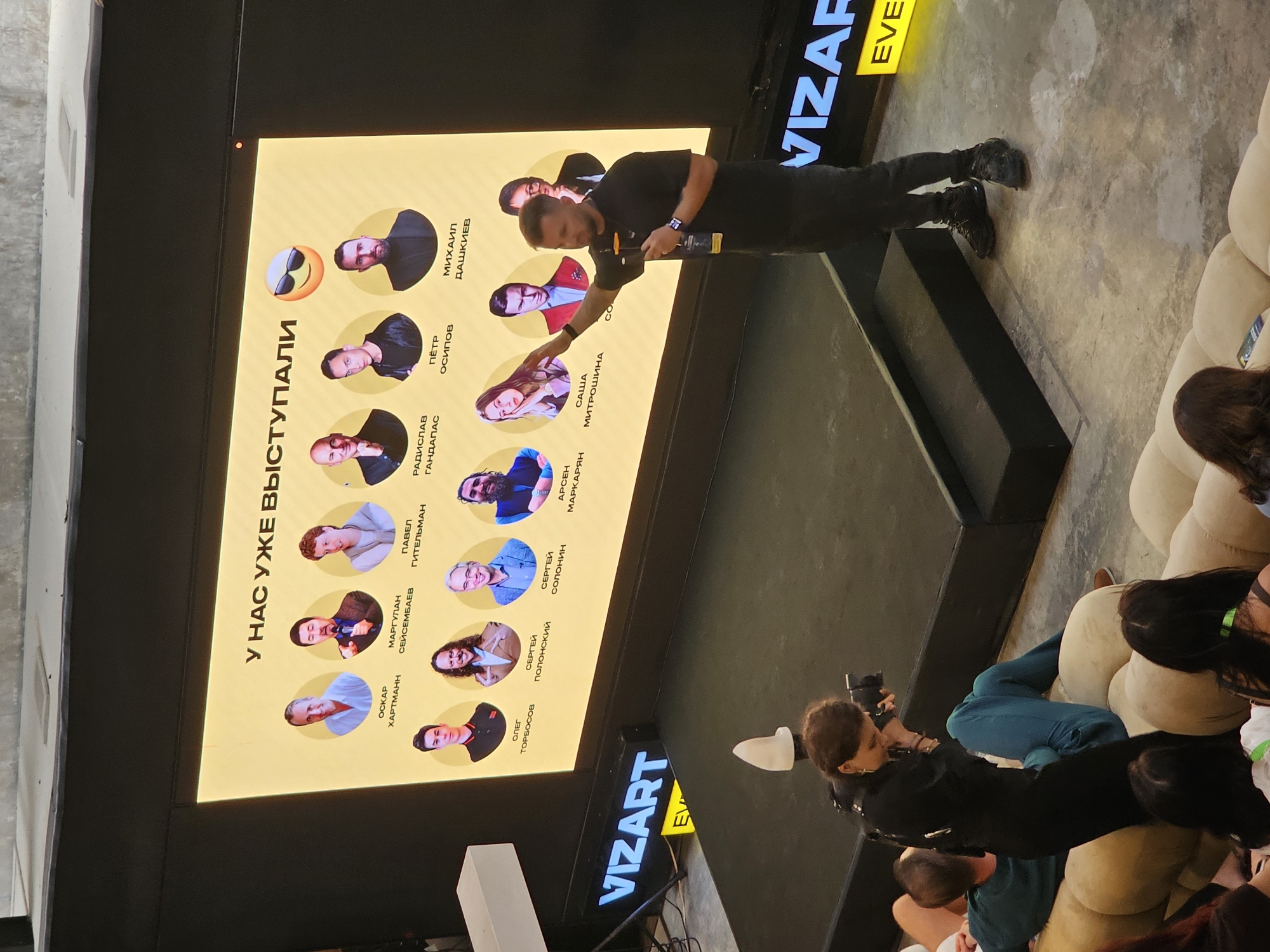 The image shows a man on a stage presenting in front of an audience. The man is standing on a slightly elevated platform. He is wearing a black shirt and pants. He is gesturing toward a large screen behind him, which displays headshots and names of people who have previously spoken. In the lower-right corner of the image, part of the audience is visible, seated on couches. In the foreground, a person is taking photos with a camera. A sign displaying the word "VIZART" is visible in the upper right and lower center of the frame. The location in North Kuta, Indonesia is not evident in the photo.