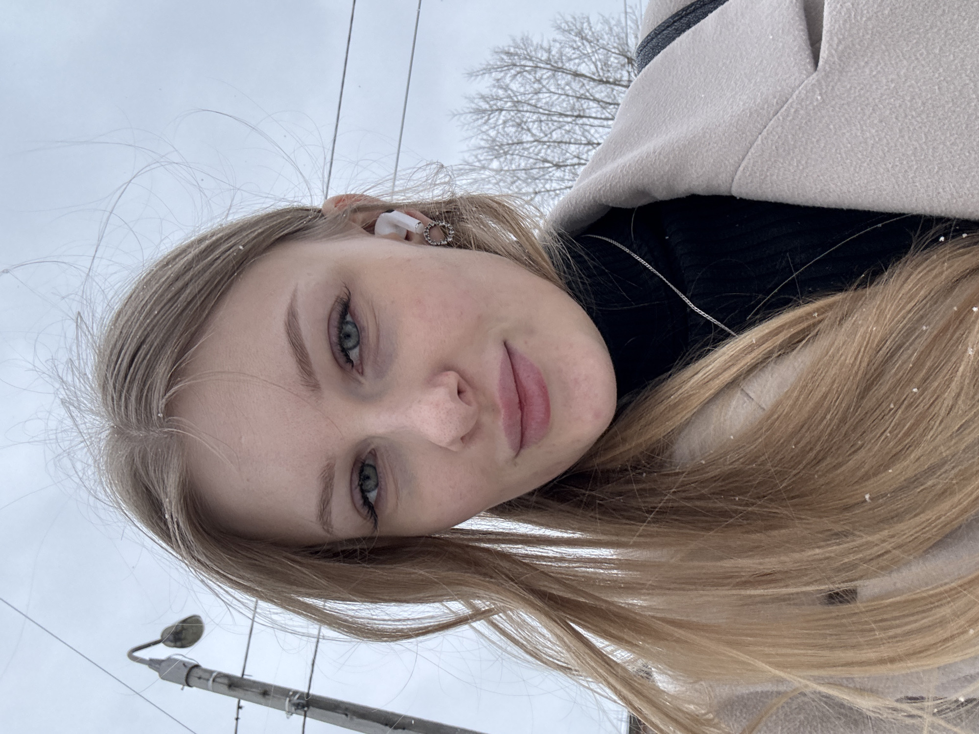 A close-up portrait of a fair-skinned young woman with long blonde hair, presumably taken in Perm, Russia based on similar images, is presented at an angle. The woman has light eyes and is wearing a black turtleneck sweater and a beige coat. She has a white earbud in her ear and a silver necklace around her neck. There are small white particles, likely snowflakes, on her hair and coat. The background is a blurred view of a grey sky and a leafless tree, with power lines and a lamppost visible in the lower left corner of the frame. The focus is primarily on the woman's face.