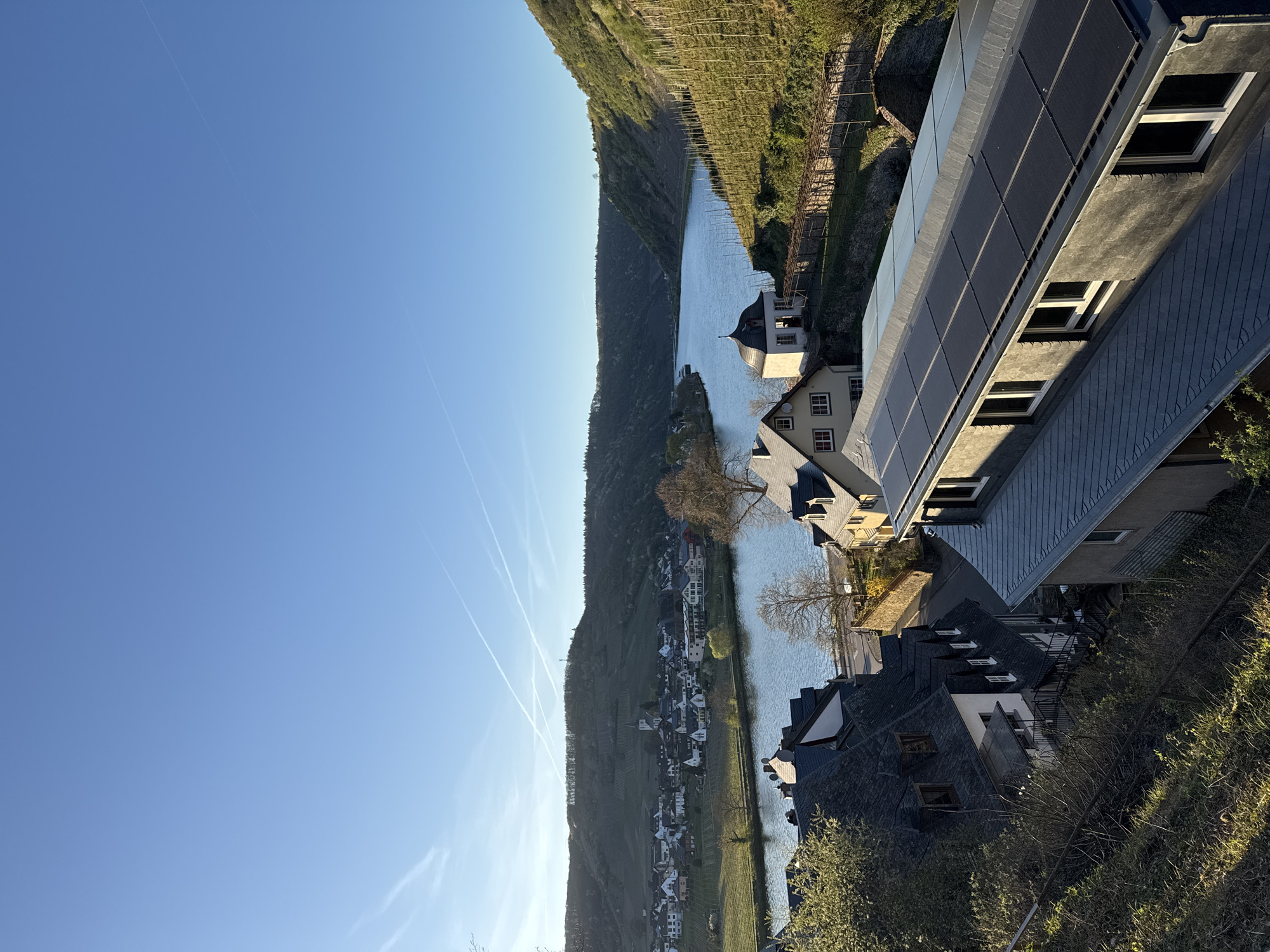 The image presents a landscape in Beilstein, Germany, featuring the Moselle River winding between steep, vineyard-covered hills. The river's surface reflects the sky, and several contrails are visible in the blue sky. Along the riverbank, closely packed houses with dark roofs, including a building with integrated solar panels, form the town. Trees with bare branches are interspersed among the buildings. The photograph appears to be taken from an elevated position.