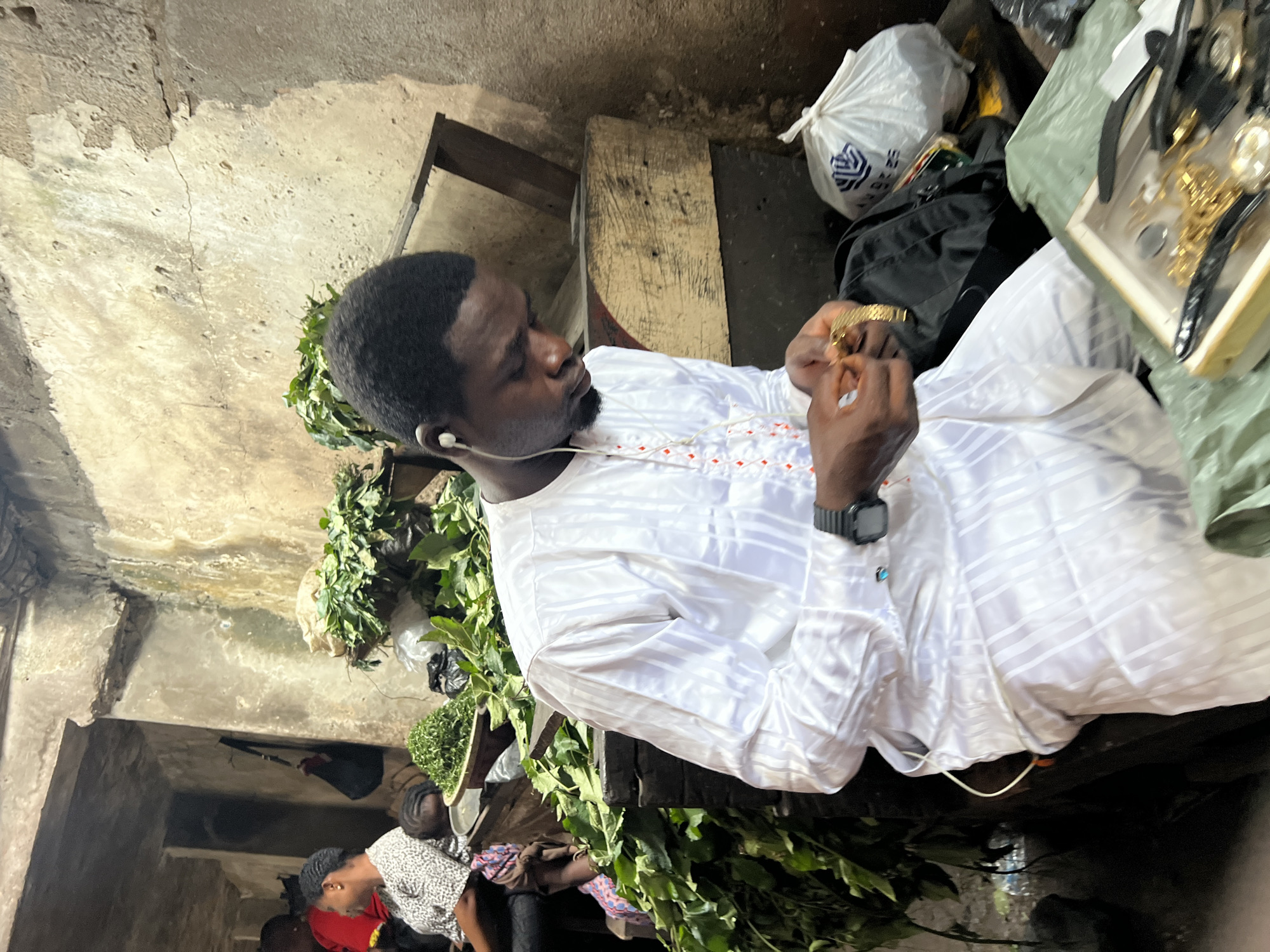 In Nkpor, Nigeria, a man wearing a white traditional outfit with orange embroidery is seated in a market stall. He has earbuds in and is focused on examining a gold-colored bracelet. To his right, a white plastic bag with a logo and a black backpack are visible. In the foreground, a wooden box contains various gold-colored jewelry, including necklaces and watches, on top of green fabric. The background shows stalls with piles of leafy green vegetables, and other people can be seen in the periphery. The environment appears to be an indoor market with rough, textured walls.