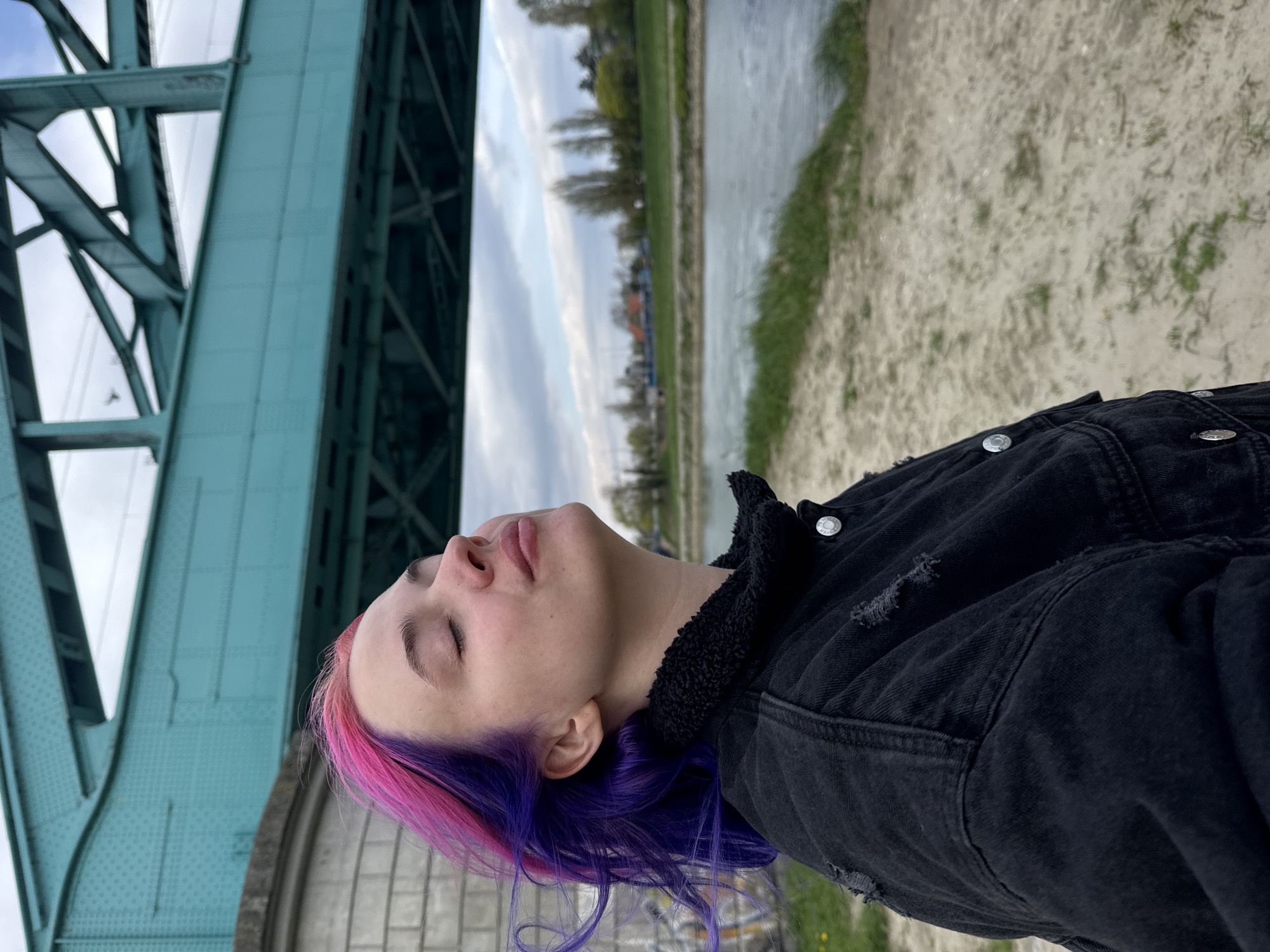 The image shows a person with pink and purple hair, wearing a black denim jacket with a sherpa collar. They are under a blue bridge, with a river and cityscape in the background. The person's eyes are closed, and their head is tilted back. The location is in the city of Zagreb, Croatia, as evidenced by the distinctive architecture of the bridge. The area appears to be a park or riverside pathway.