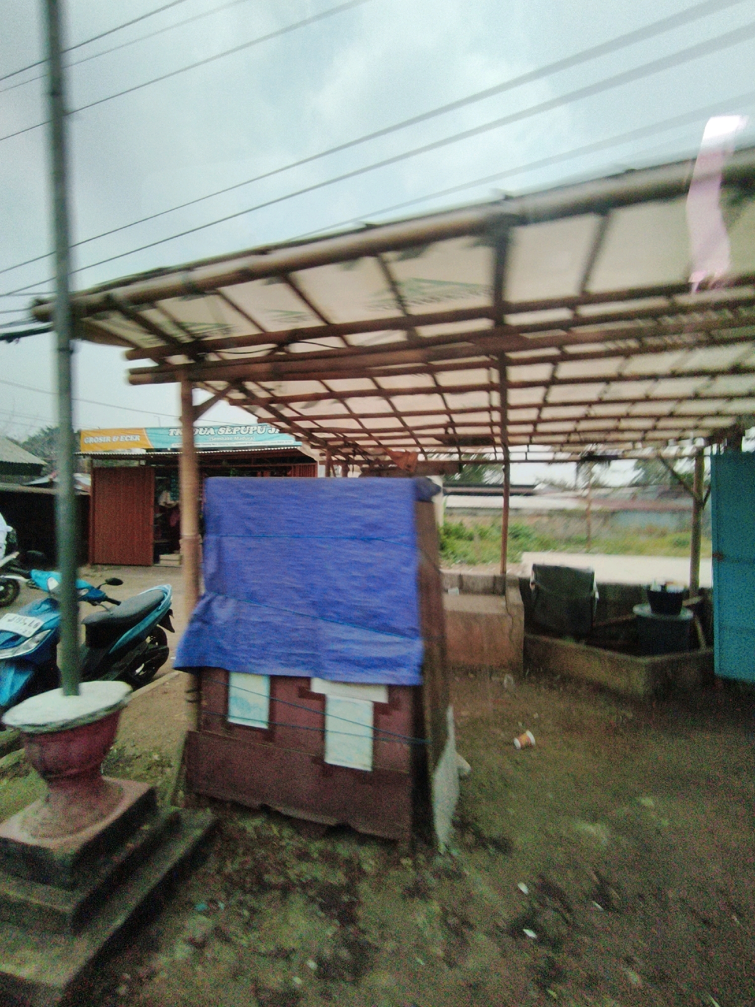 The image depicts an outdoor scene in Sukaraja, Indonesia. In the foreground, a small, makeshift structure is covered with a blue tarp. To the left, a turquoise motorcycle is parked. Behind the motorcycle, a red door is visible, part of a building with a sign that reads "GROSIR & ECER" and "TK dua sepupuu." A bamboo-covered canopy extends over the scene. In the background, a field with some vegetation and a fence can be seen. The sky is overcast. The overall impression is of a utilitarian area, possibly a marketplace or roadside stall.