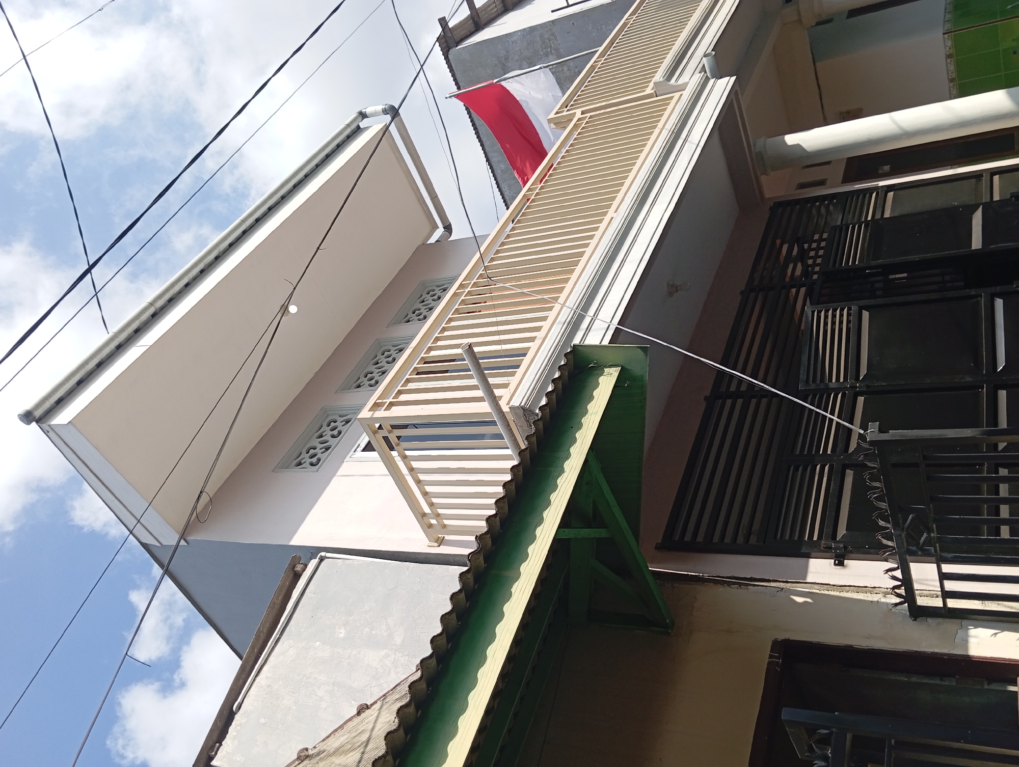 A low-angle, full outdoor shot captures the exterior of a two-story building under a bright, partly cloudy sky. The building is predominantly light pink with a white overhang. A beige balcony railing runs across the second story, and behind it, an Indonesian flag is mounted on a pole. The first story features a black metal gate with spiked finials, and a dark-colored door. A green corrugated metal awning extends from an adjacent structure. Overhead, a network of black electrical wires is visible against the sky. The scene appears to be a residential area.