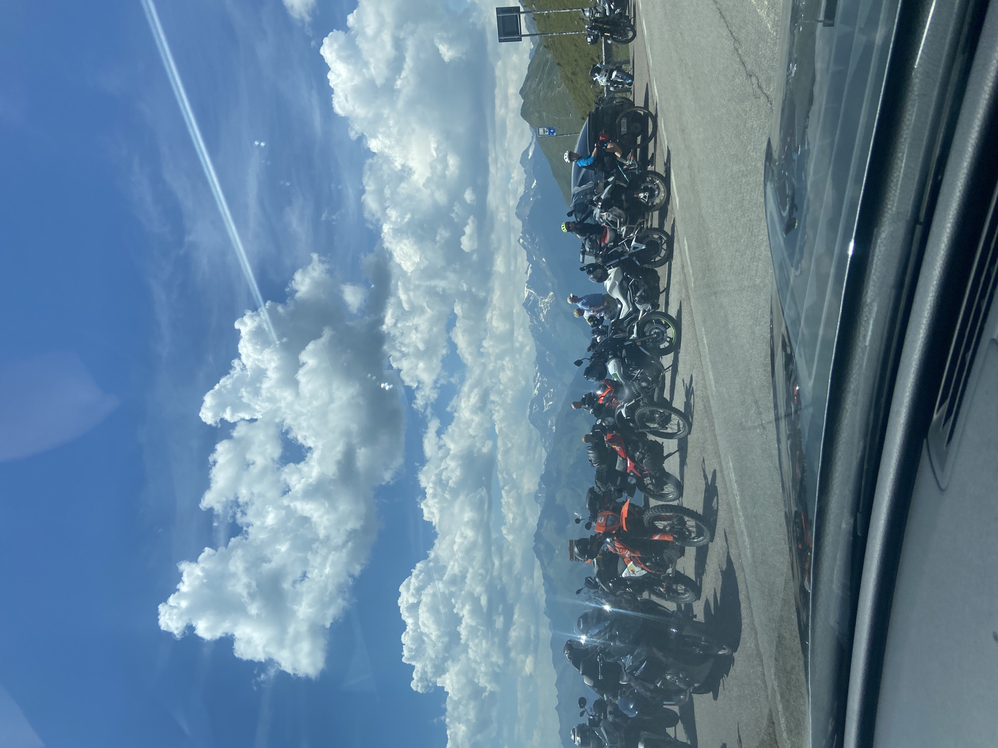 The image captures a scenic view of a mountain pass, likely in the Italian Alps, with a group of motorcycles parked by the roadside. In the foreground, the dashboard of a vehicle suggests the photo was taken from inside a car. The main subjects are a collection of dual-sport and adventure motorcycles, varying in color, parked in a line along the asphalt road. Some motorcycles are orange, red, black, and white. Several individuals are visible, some standing near the bikes, and others are seated on them, wearing helmets and motorcycle gear. The backdrop features majestic snow-capped mountains under a bright blue sky adorned with fluffy white clouds. The environment appears to be a high-altitude location, possibly a popular stop for motorcyclists. A signpost can be seen on the right side of the image, and a few more signs further up the hill, suggesting it is a marked viewpoint or pass. The overall scene conveys a sense of adventure and appreciation for the mountainous landscape. The location is identified as St. Leonhard in Passeier - San Leonardo in Passiria, Italy.