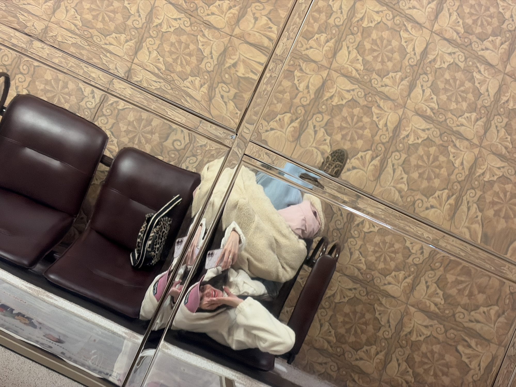 The image, taken from a high, overhead angle, captures a reflection on a highly polished or mirrored surface, which is divided by horizontal and vertical strips. This reflective surface reveals an intricate, light brown and beige geometric pattern, which also serves as the direct background, suggesting tiled flooring or wall panels adorned with floral and swirling motifs.
In the prominent reflection, a person is seen reclining on dark brown public seating, resembling leather or faux leather, parts of which are also directly visible in the foreground. The individual is wearing a cream-colored, fluffy jacket or coat, a light blue top, and a pink baseball cap. They are holding a smartphone, possibly an iPhone, and appear to be taking a selfie or engaging with the device, displaying a cheerful, smiling expression. One foot, clad in a patterned (potentially leopard print) shoe, and another in a white slipper with pink accents, are visible in the reflection. A small leopard print bag with a black and white striped strap rests on an adjacent seat.
The scene depicts a moment of relaxation in a public indoor environment, likely a waiting area in Saint Petersburg, Russia, such as an airport, train station, or a similar facility. The person's casual attire and happy demeanor, combined with their use of the smartphone, suggest a leisurely activity. There is no visible text in the image.