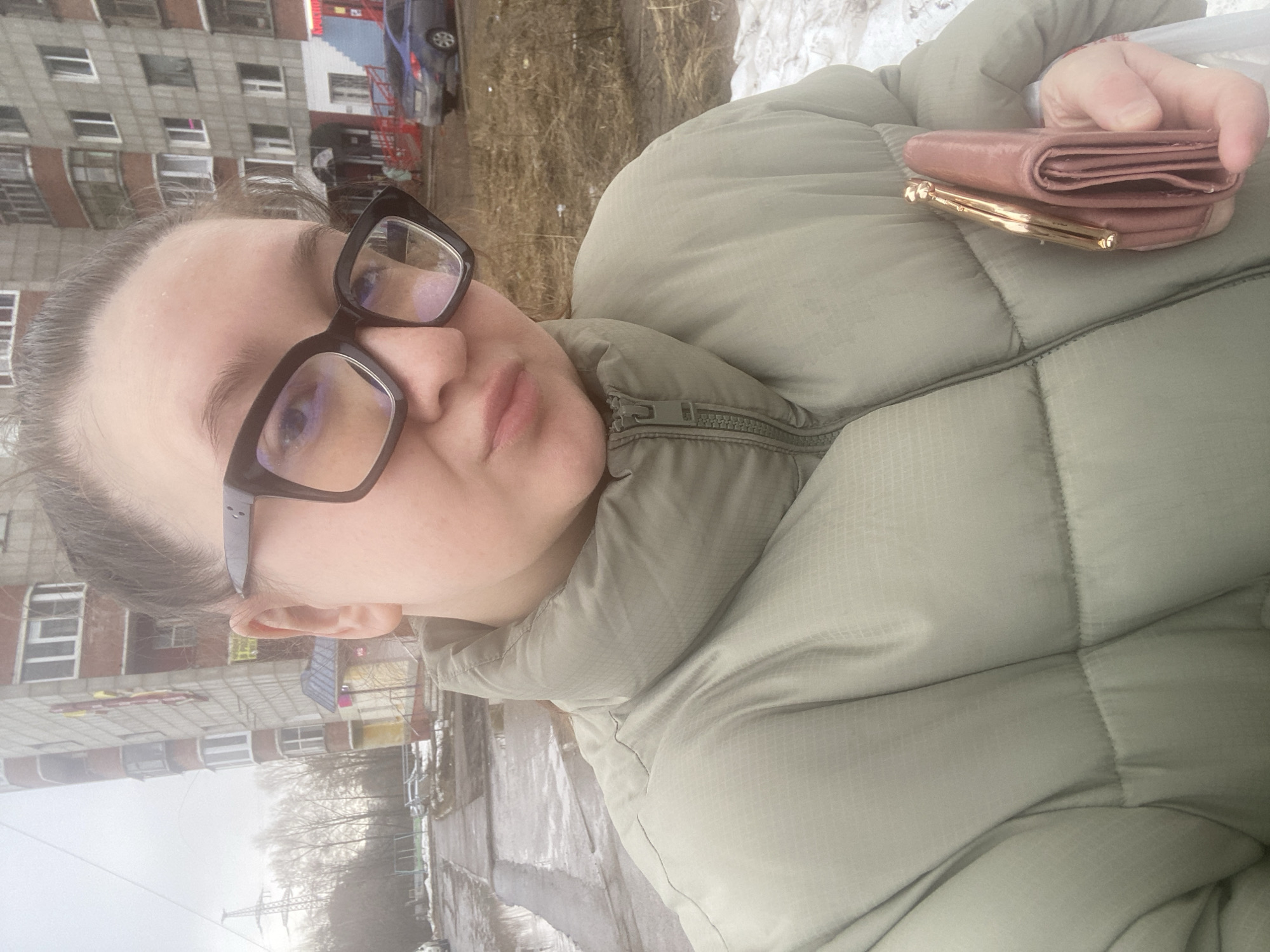 A person wearing glasses and a puffy jacket holds a wallet in their hand, with a residential building and trees visible in the background. The location is Chaikovsky, Russia, as indicated by the architectural style of the building and the urban environment. The ground is wet, suggesting recent rain or snow melt.