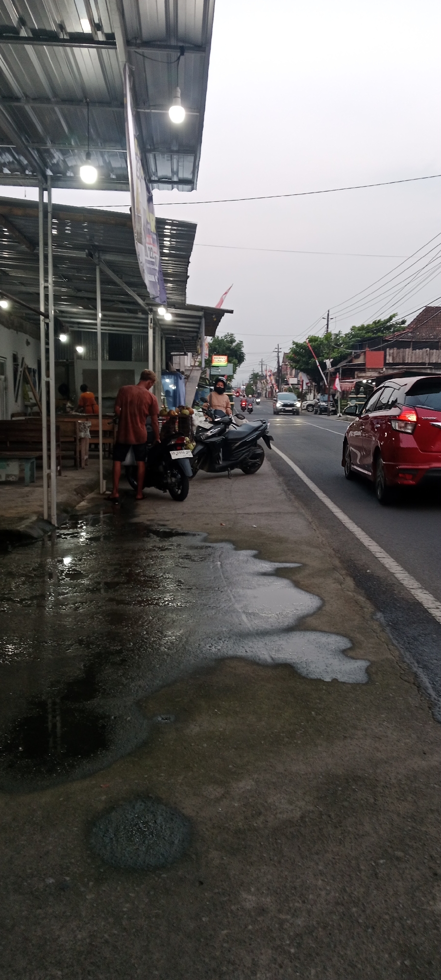 This image depicts a street scene in Maguwoharjo, Indonesia. In the foreground, a man in an orange t-shirt and shorts stands by a parked motorcycle on a wet sidewalk. To his left, a covered food stall with metallic roofing is visible, with a few people inside. In the background, the street is lined with buildings and trees. Traffic, including cars and motorcycles, moves along the road. The sky is overcast. The overall impression is of a roadside establishment in a somewhat humid or recently rained-on environment.