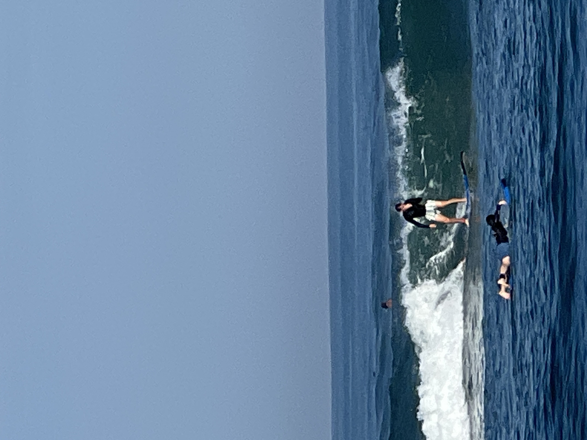 In the ocean off the coast of North Kuta, Indonesia, a surfer is riding a wave. The surfer, dressed in a black long-sleeved rashguard and white shorts, stands on a blue surfboard, balancing on the breaking wave. Behind the surfer, another person is visible in the water, further out in the ocean. In the foreground, two individuals are lying on their backs in the water, one on a blue flotation device, appearing to be resting or observing. The water is a deep blue, with white foam from the breaking waves. The sky is a clear, light blue.