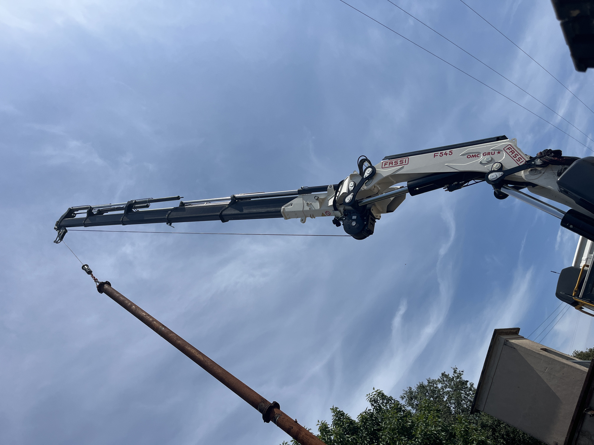 An outdoor, low-angle, medium shot captures a construction scene against a blue, partly cloudy sky in Castiglione delle Stiviere, Italy. The primary focus is on a white and black FASSI F54-5 truck-mounted crane's articulated arm, which is extended upwards. The crane's branding, "FASSI" and "OMC GRU," is visible on its white segments. A thick, rusty, brown pipe is suspended from the crane by a cable and a metal clamp. The pipe angles diagonally from the left to the center of the frame. In the lower left, the green foliage of a tree and the upper edge of a small, light brown, boxy structure are visible. In the lower right corner, a portion of a white vehicle, possibly the truck on which the crane is mounted, is seen, along with a few dark overhead power lines. No people are visible in the image. The scene appears to depict a pipe being lifted or moved by the crane.