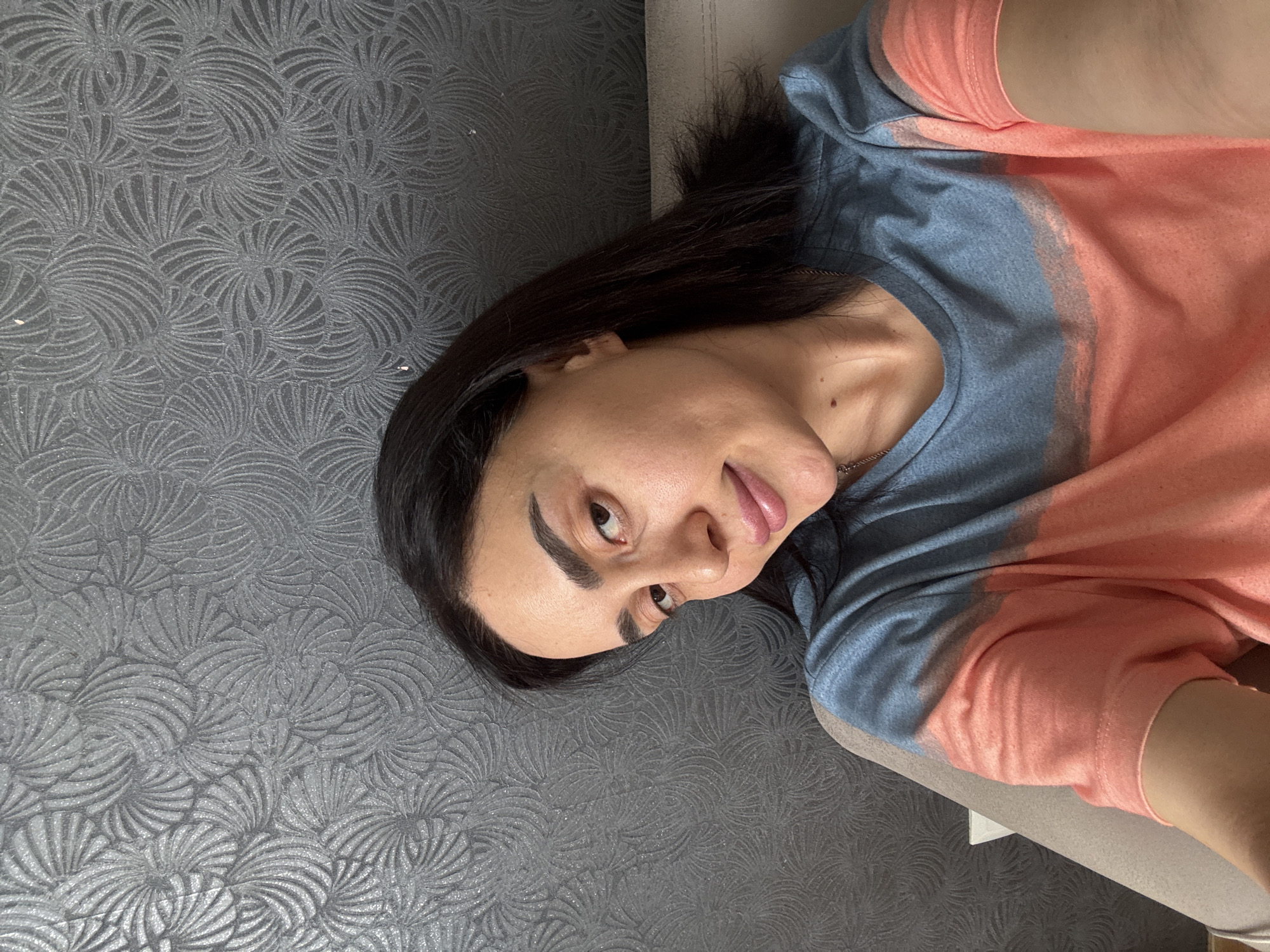 A woman is captured in a close-up portrait, presumably indoors in Tbilisi, Georgia. She appears to be reclining on a light beige or grey upholstered surface, likely a sofa or bed. Her head is positioned towards the left of the frame, and she is looking directly at the camera with a playful expression, her tongue slightly out. She has long, dark hair and prominent, well-groomed dark eyebrows framing her dark eyes.
She is wearing a distinctive two-toned t-shirt that transitions from a muted blue-grey at the top to a vibrant peach or coral color towards the bottom, suggestive of a tie-dye or ombre design. A thin, silver-colored chain necklace is visible around her neck, and a small mole can be seen on her upper chest or collarbone area.
The background features a dark grey wall covered in a richly textured wallpaper. The pattern is intricate, consisting of repeating fan-like or stylized leaf motifs that appear to have a slight sheen, contributing to the domestic setting. There is no visible text in the image.