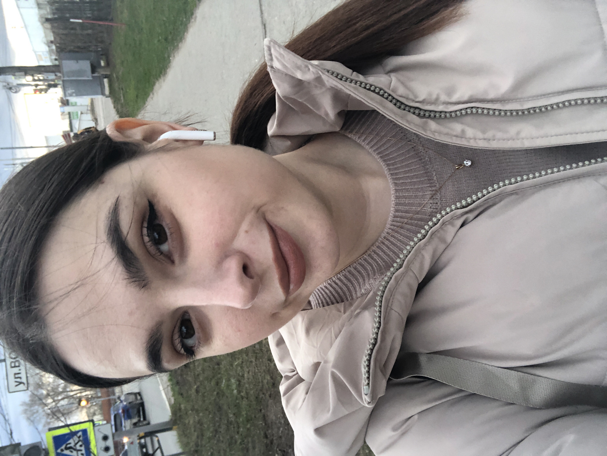 The image is a vertical, close-up selfie of a woman outdoors, captured at a slightly tilted angle. The woman, with dark brown hair and dark eyes, looks directly at the camera with a neutral to subtly smiling expression. She is wearing a light beige or tan padded jacket, zipped up, over a ribbed knit sweater in a similar muted brownish-beige tone. A small, sparkly pendant on a thin chain is visible at her neckline. A white wireless earbud (resembling an AirPod) is placed in her right ear.
The background, largely out of focus, indicates an outdoor urban or suburban setting. To her left, a sidewalk or path is visible alongside an area of green grass and some bare trees, suggesting a cooler season. Utility poles, distant buildings, and what appears to be a road or street are present further back. In the lower left corner, a partially visible street sign displays Cyrillic text, "ул. Во...", likely indicating a street name, and a blue and white pedestrian crossing sign can also be seen. The lighting is soft and diffused, possibly from an overcast sky or during the late afternoon. The scene conveys a casual moment, perhaps while she is out walking in Chaikovsky, Russia.