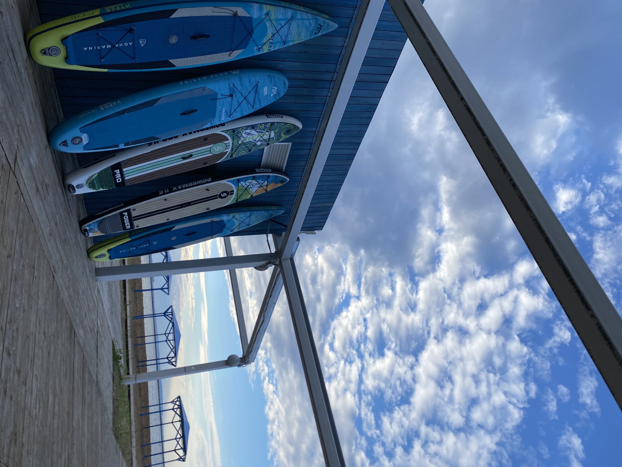 The image captures an outdoor scene with several stand-up paddleboards leaning against a blue wooden wall. The paddleboards, varying in color and design, are arranged in a row. Above the wall, a metal structure extends, supporting a white canopy or awning. The background reveals a body of water, possibly a river or lake, with several blue canopy structures along the shoreline. The sky is partly cloudy, with a mix of blue sky and white clouds, suggesting either early morning or late afternoon. The ground in the foreground is made of wooden planks. This scene is located on the embankment of the Kama River in the city of Chaikovsky, Russia.