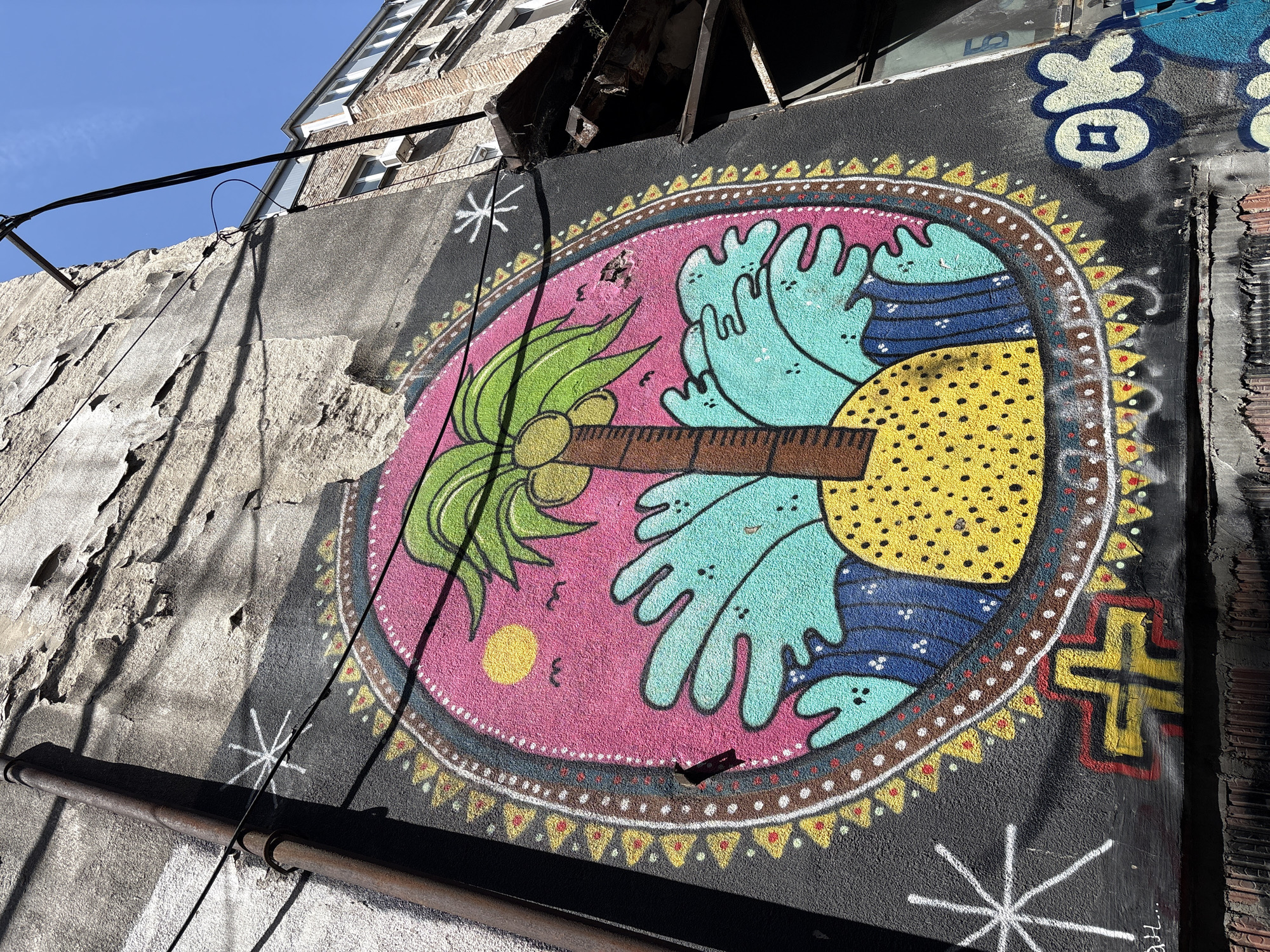 An exterior shot of a mural on a weathered gray wall, presumably in Belgrade, Serbia. The mural is centered and oval-shaped, framed by a decorative border of dots and geometric shapes in brown, yellow, white, and red. The oval's interior is divided into a pink background featuring a yellow circle, a group of stylized light-blue hand-like forms, and a yellow textured circle containing blue wavy lines. At the left, a palm tree with green leaves and brown coconuts extends from the center toward the edge of the oval. The wall is cracked and chipped, especially on the left, where the exposed brick shows through. To the right of the mural, a section of stacked brick is visible. The gray wall also includes other smaller graffiti elements: A white star-like figure below the main design, a blue and white symbol above, and a yellow and red cross-like shape to the right. A rusted pipe crosses the lower left portion of the wall. Above the mural, the facade of a multi-story building is seen against a clear blue sky. Wires stretch across the top left of the frame.