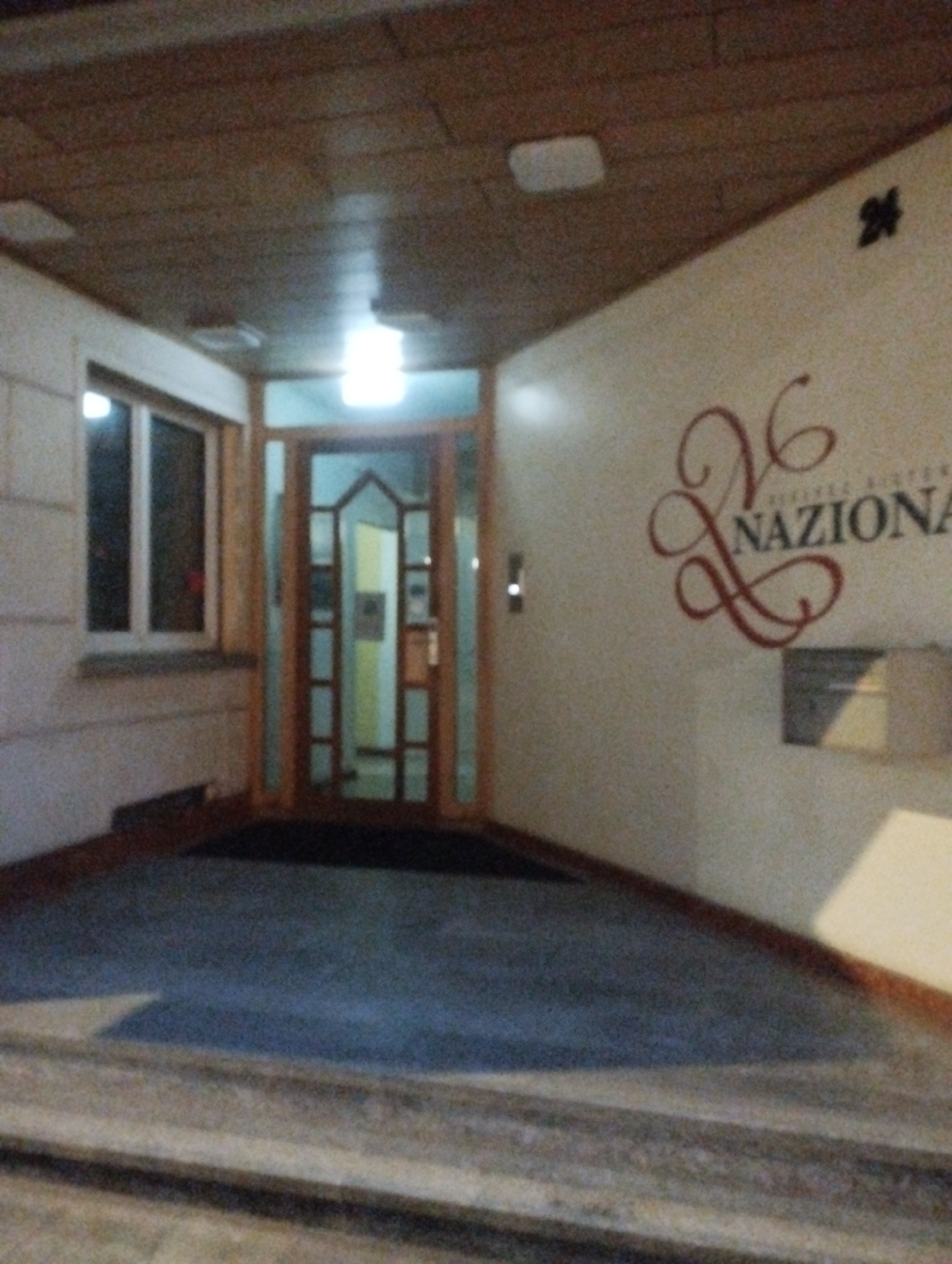 The image depicts the exterior entrance of a building at night. The entrance features a double glass door with a wooden frame, leading into a brightly lit interior hallway. To the left of the entrance is a large window with a white frame, also showing interior light. The wall to the right of the entrance is light-colored and features a decorative logo with the number "26" and the word "NAZIONA". Above the entrance, the ceiling is made of dark wood panels. A number "24" is visible on the upper right exterior wall. The ground in front of the entrance is paved with dark material, and a light-colored step is visible in the foreground. The overall impression is of an apartment building or a commercial establishment.