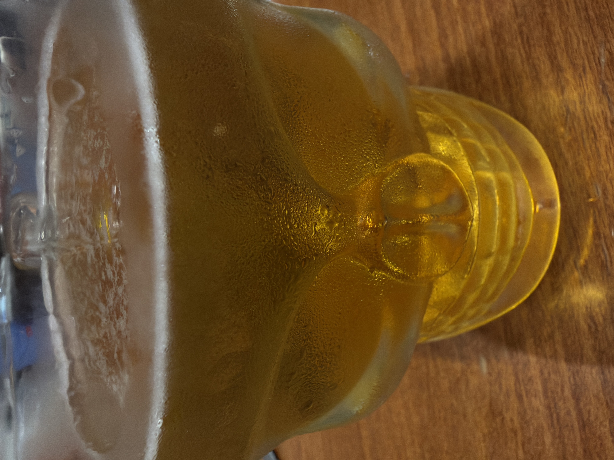 A close-up shot shows a skull-shaped glass filled with a golden-colored beverage, likely beer, with a frothy head. Condensation beads are visible on the exterior of the glass, indicating it is chilled. The glass rests on a wooden surface, possibly a table. The background is blurred but suggests an indoor setting. No people or specific city locations are identifiable within the image itself.