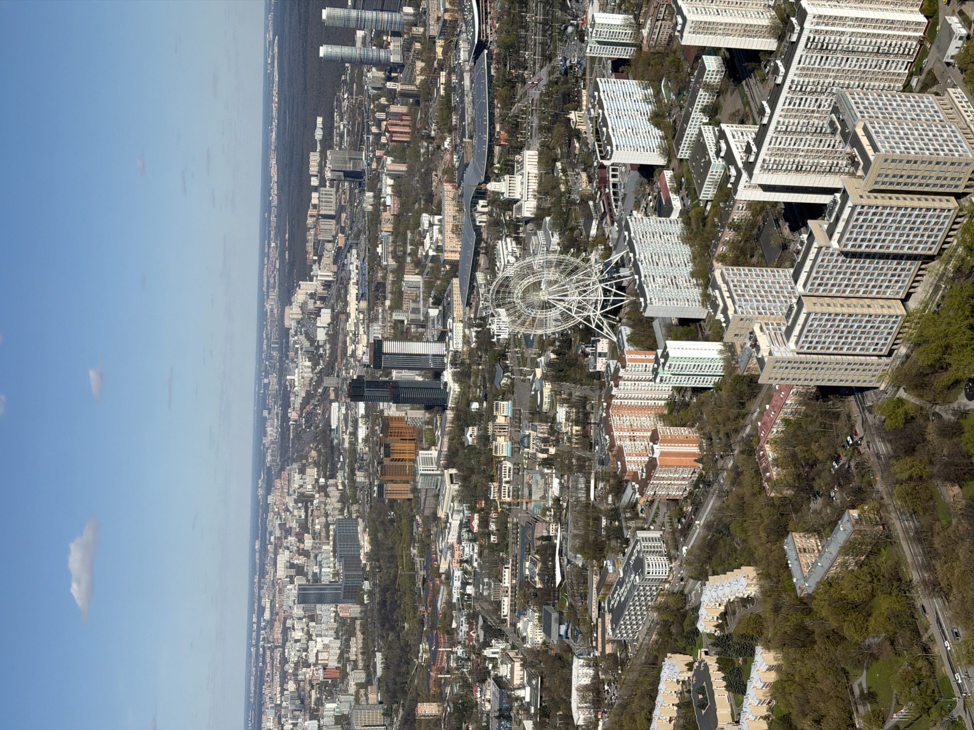 The image shows a high-angle view of a city, with a clear blue sky visible to the left and a dense forest edge in the distance. The city is densely populated with a variety of buildings, including high-rise apartments, commercial structures, and what appear to be governmental or institutional buildings. A distinctive Ferris wheel stands out prominently in the middle ground. The buildings are arranged along an urban landscape. The scene is consistent with an urban environment on a bright day. The image does not provide enough information to confirm the exact location.