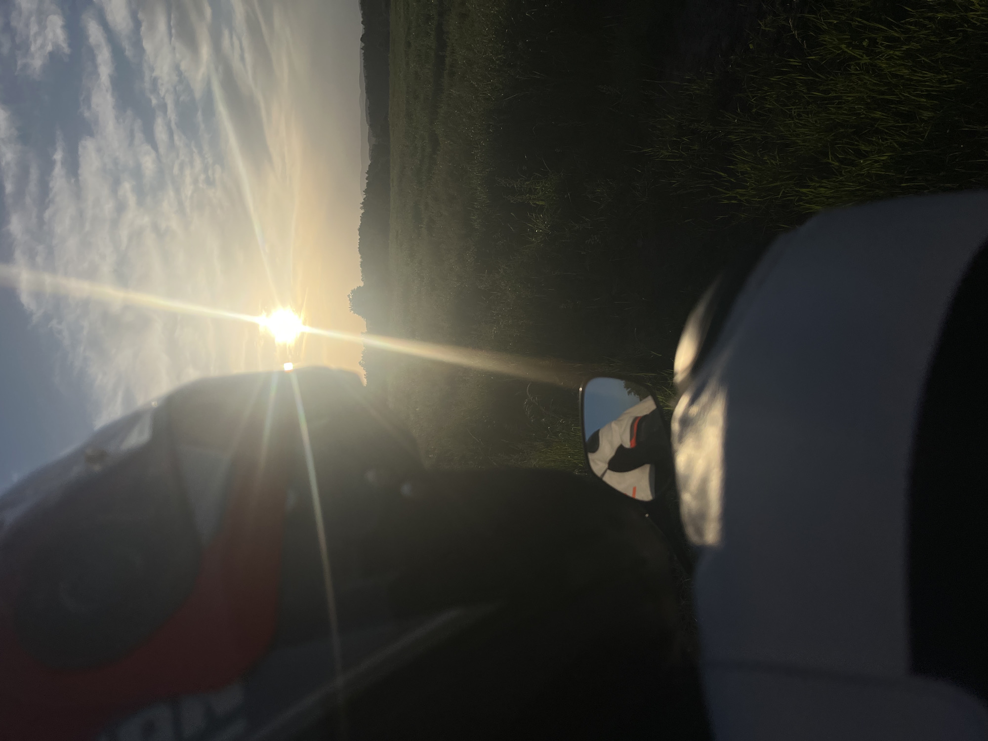 The image captures a view from what appears to be a motorcycle, with the sun setting in the background. The sky is visible with clouds and bright sunlight. A field or area of dense vegetation is present to the right of the sun. A motorcycle helmet, with a red and white design, is partially visible in the foreground on the left. The motorcycle's side mirror is visible, reflecting a rider wearing a white jacket. The location is unidentifiable.