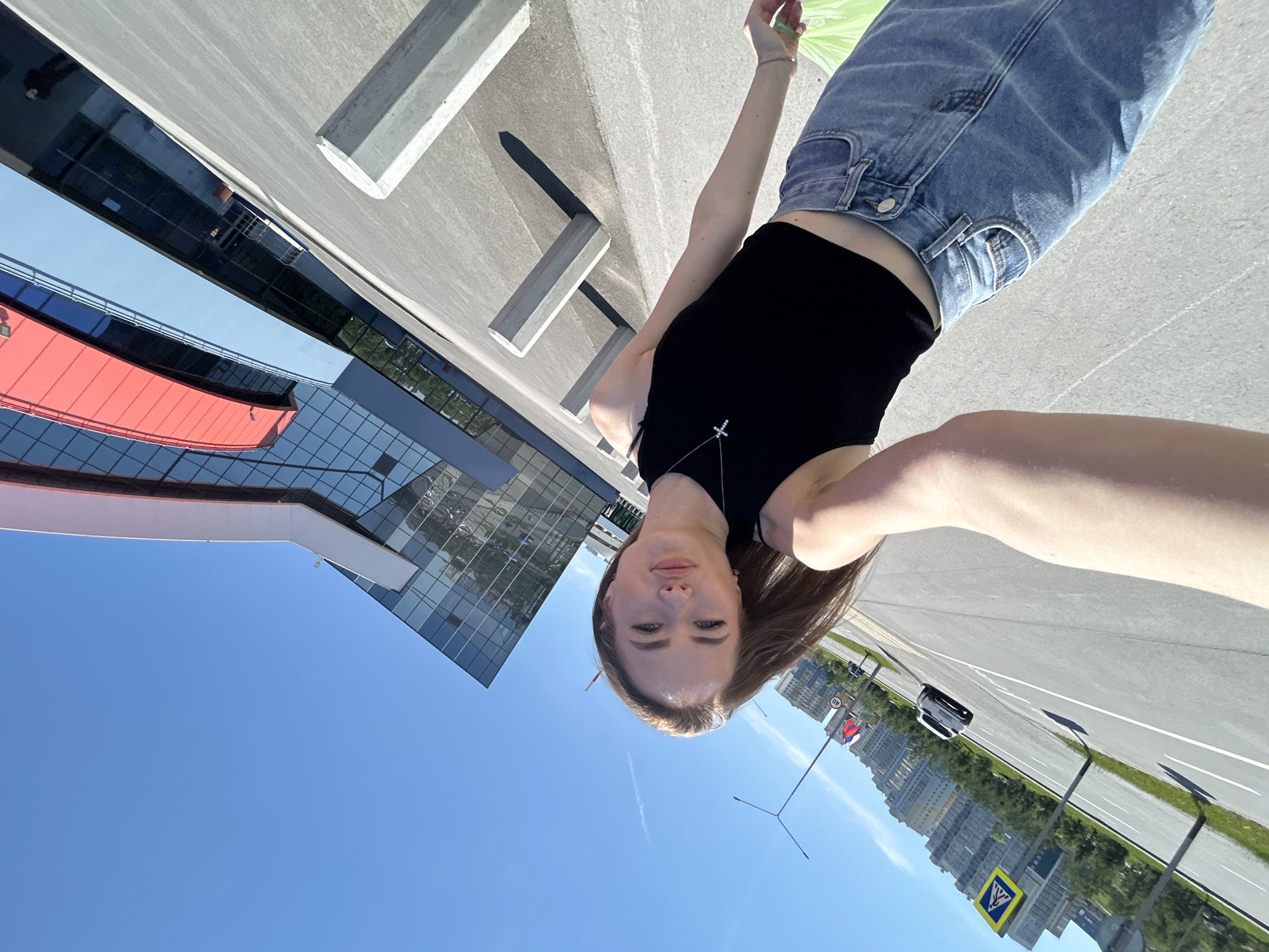 The image is a tilted, eye-level selfie featuring a young adult female outdoors on a bright, sunny day. The woman, with fair skin and light brown hair, is positioned in the foreground, facing the camera with a neutral expression. She wears a black sleeveless crop top, light blue jeans, and a silver-toned cross necklace. Her right arm is extended, holding the camera, with a light green plastic bag partially visible in her right hand.
Behind her, to the right, is a large, modern building featuring a predominantly glass facade that reflects the blue sky and surrounding environment. Sections of the building are accented with red and light blue horizontal paneling. To the left and in the mid-ground, a multi-lane asphalt road with white markings (including a double solid line and a broken line) stretches into the distance. A circular white "40 km/h" speed limit sign with a red border and a blue and yellow pedestrian crossing sign are visible alongside the road.
Several grey concrete bollards or car stops are arranged on the paved area next to the building, casting distinct, elongated shadows to the left, indicating direct sunlight from the right. Further along the road, a white car is visible, along with multiple streetlights and green foliage. In the far background, a cluster of multi-story residential buildings is visible under a clear blue sky, which features a faint white contrail. A Russian flag (white, blue, red horizontal stripes) is prominently displayed on a pole by the roadside. The overall scene depicts an urban or suburban setting with modern architecture and infrastructure.