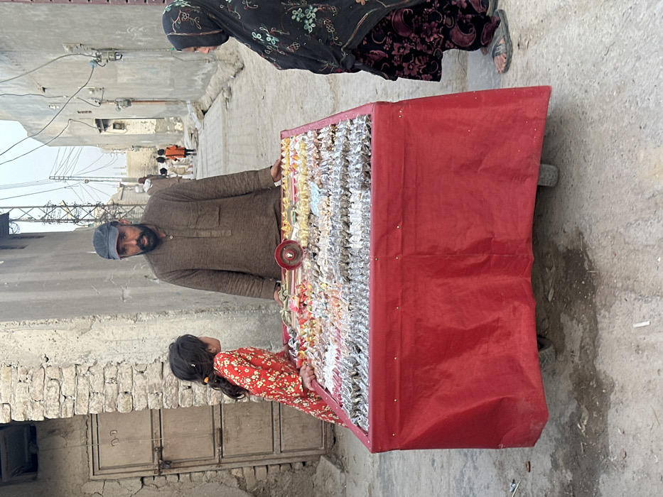 A street scene in Quetta City Tehsil, Pakistan, depicts a male vendor operating a mobile merchandise cart, attended by two individuals. The vendor, an adult male with a dark beard and cap, stands centered behind a rectangular cart draped in red fabric. His hands rest on the cart's surface, which is laden with multiple rows of assorted small items, including what appear to be bangles, trinkets, or candies, arranged for display. To the vendor's left, a young girl in a red floral dress leans against the cart, looking towards the vendor. To the vendor's right, an individual wearing a dark embroidered headscarf and patterned clothing stands near the cart. The environment consists of a narrow, unpaved dirt street flanked by utilitarian, light-colored plastered buildings. Overhead power lines and a metal utility pole are visible in the background. In the distance down the street, several additional figures, including children, are visible walking away from the viewer. The lighting suggests daylight conditions.