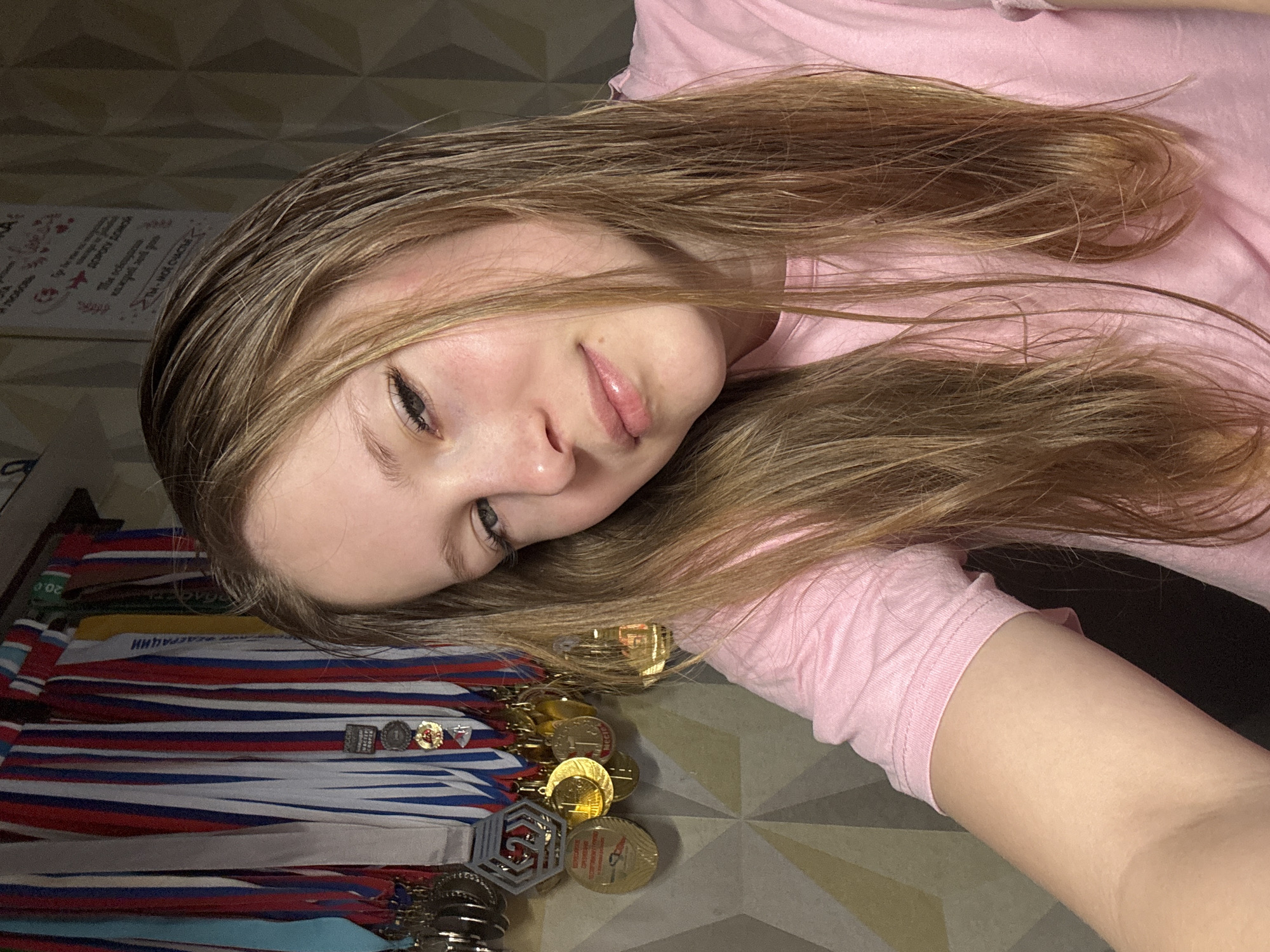A young person with light brown hair and wearing a pink shirt is lying on their side. They are surrounded by medals, some with ribbons in the colors of the Russian flag. The location is possibly in the city of Chaikovsky, Russia, based on the flag colors, but without further information, this is speculative. The individual is looking at the camera with a slight smile. The background appears to be a patterned surface.