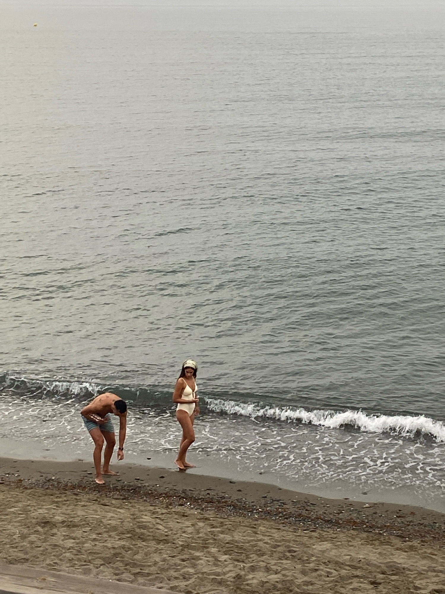 The image depicts a sandy beach in Estepona, Spain, with the gentle waves of the Mediterranean Sea lapping at the shore. A man, wearing blue striped swim shorts, is bent over on the wet sand, appearing to examine something on the ground. Nearby, a woman in a white swimsuit and a headband stands facing the sea. The water is a muted gray-blue, with white foam from the breaking waves. The sky is overcast, contributing to the diffused light. The beach is composed of sand interspersed with small pebbles and shells.