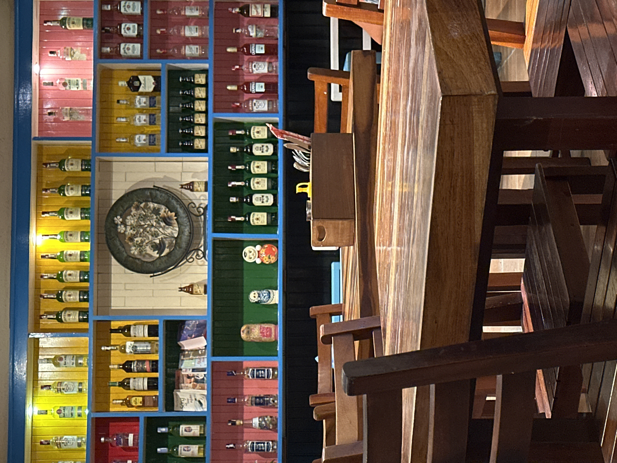 The image depicts the interior of a bar or restaurant in Nha Trang, Vietnam, characterized by brightly colored shelves filled with an assortment of liquor bottles. The shelves are organized into distinct sections with vibrant backdrops of yellow, pink, and blue. On the topmost shelves, various bottles of clear spirits and liquors are displayed, including what appears to be Bacardi and Jameson. Below these, more shelves showcase different brands of alcohol, such as vodka and rum, interspersed with what seem to be wine bottles.
On a middle shelf, a decorative round plate with a floral motif is prominently displayed, flanked by small bottles and what might be ceramic figurines, including a matryoshka doll and a round, red-faced doll. Further down, more rows of bottles continue the theme of a well-stocked bar.
In the foreground, several wooden tables and chairs are arranged. One table in the center has a wooden box containing cutlery and what looks like a red marker. The lighting appears to be a mix of ambient room lighting and spotlights from above, highlighting the bottles and the polished wooden surfaces of the tables and chairs. The overall atmosphere suggests a lively establishment, possibly a popular spot for drinks and socializing in Nha Trang.