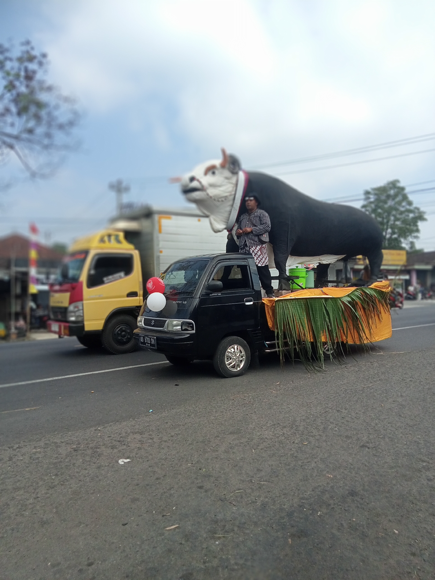 In the city of Urutsewu, Indonesia, a black Suzuki Carry pickup truck is parked on the right side of a paved road. The truck's bed is decorated with an orange cloth and large, green palm fronds. Perched on top of the truck's bed, and extending over the cab, is a large, black inflatable bull with a white head and red and white trim around its neck. A person wearing a striped shirt, dark pants, and a patterned sarong stands on the truck bed, next to the bull. Red and white balloons are attached to the front of the truck's cab. In the background, a yellow and red truck is parked behind the pickup truck. Buildings, trees, and a utility pole are visible further in the background, suggesting a semi-urban or rural roadside environment. The sky is overcast with some blue patches. The overall scene depicts a procession or parade float.