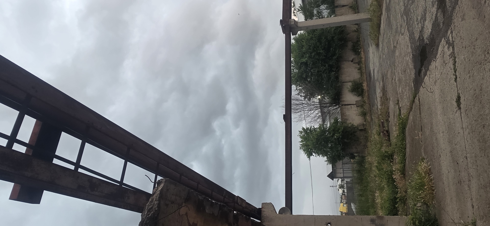 This image depicts an outdoor scene characterized by industrial decay and an overcast sky. The shot is composed with a wide, grey, cloudy sky occupying the upper portion of the frame. The clouds appear thick and uniformly grey, indicating a cloudy day without direct sunlight.
In the left foreground, a substantial, dark, and rusty metal structure, resembling a large beam or girder with a skeletal or truss-like design, stretches across the upper left. Below this, a portion of a concrete or stone wall is visible.
On the right side, a tall, vertical concrete pillar stands, supporting a horizontal concrete beam. Beyond these structures, a line of trees and bushes with both green foliage and bare branches marks a boundary. The ground in the foreground and midground is a rough, cracked concrete or asphalt surface, heavily overgrown with patches of green and dry weeds and grass. Dark stains are visible on the paved surface. In the distant background, faint outlines of buildings and possibly power lines can be seen, with a small yellow object discernible towards the bottom right.
The scene conveys a sense of desolation and abandonment, typical of disused industrial or urban fringes. No people or activities are visible. The overall mood is quiet and somber, primarily due to the diffused light from the heavily clouded sky. The location is consistent with an overlooked area in Karnobat, Bulgaria. No visible text is present.