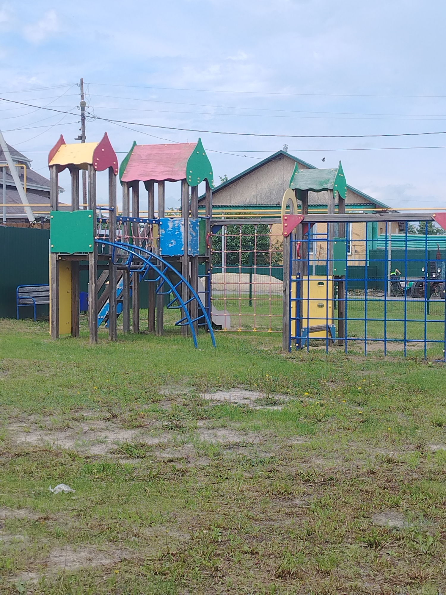 The image depicts a children's playground located in the city of Klyuchi, Russia. The playground features several colorful structures made of wood and metal, including slides, climbing frames, and platforms with pitched roofs. The ground is covered in grass and some bare patches of earth. In the background, there are houses with pitched roofs, a green fence, and power lines overhead. The sky is overcast with clouds. No people are visible in the image.