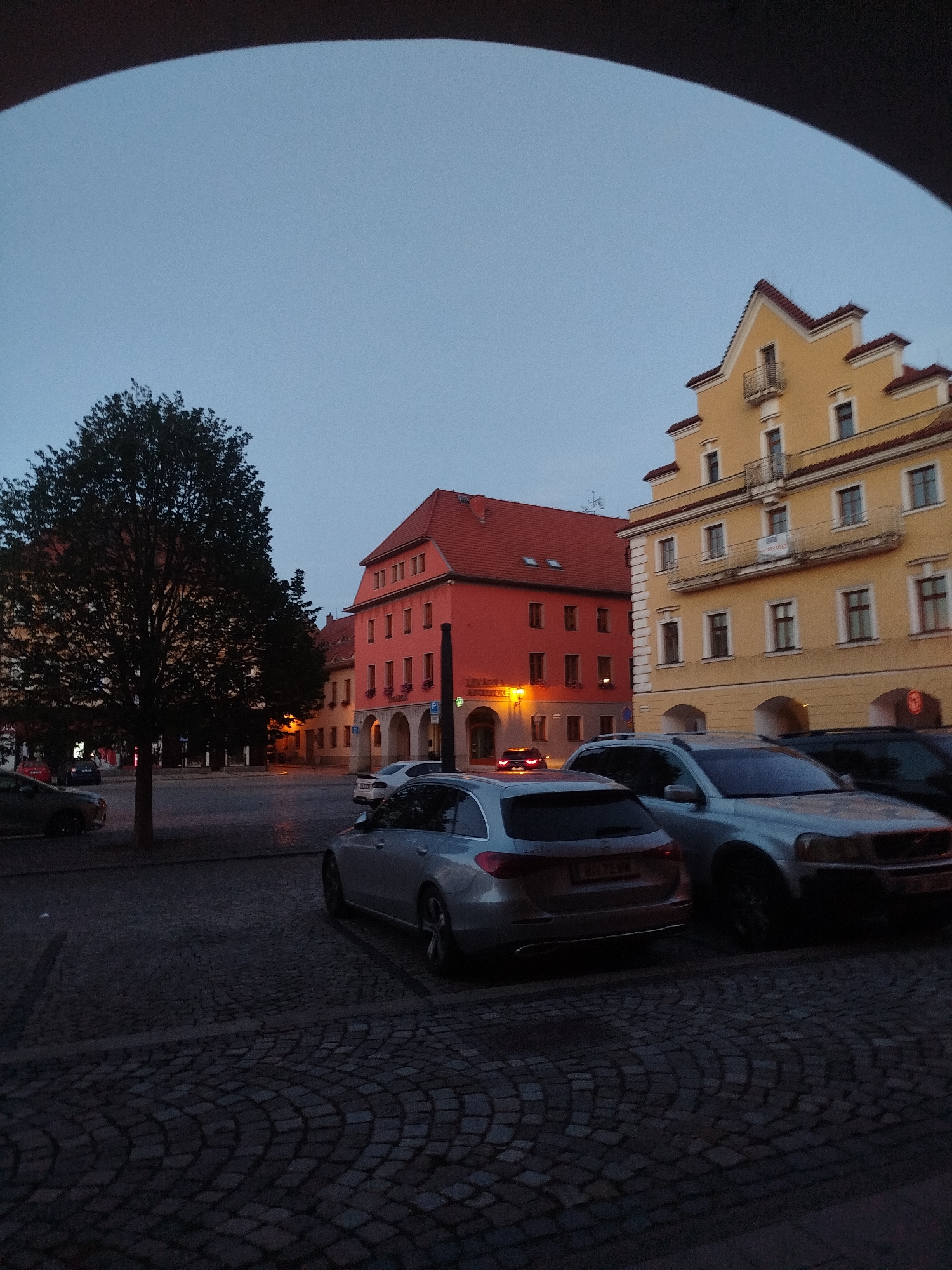 The image depicts a town square in the city of Unknown, Unknown, at dusk. In the foreground, cobblestone pavement is visible. A silver hatchback car is parked on the right, facing away from the camera, and a silver SUV is parked next to it. Behind these cars, a street separates them from buildings.
The buildings visible include a brightly lit orange building with arched doorways on the ground floor and windows on upper levels, featuring a red tiled roof. To its left, another building with a red tiled roof is partially visible, with a large tree in front of it. On the far right, a yellow building with a stepped gable roof and balconies is prominent. The sky is a dusky blue, indicating the transition from day to night. The scene appears to be quiet, with no people clearly visible.