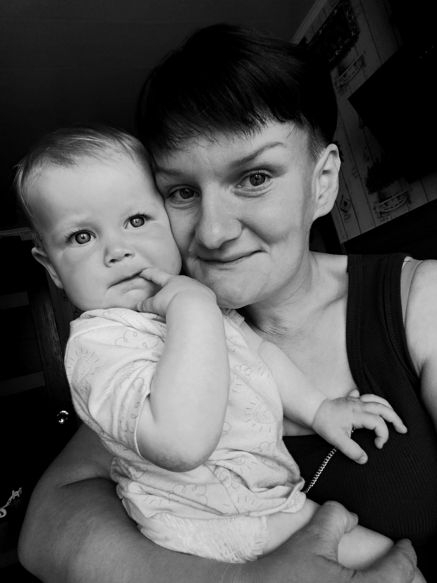 A black and white, close-up, high-angle selfie shows a woman holding a baby. The woman has short, dark hair and is looking at the camera with a slight smile. The baby, positioned in front of the woman, is looking at the camera with its mouth slightly open and a finger in its mouth. The background is dark and appears to be an indoor setting, possibly a home. There is no discernible information about the location in the city of Fokino, Russia, within the image itself.