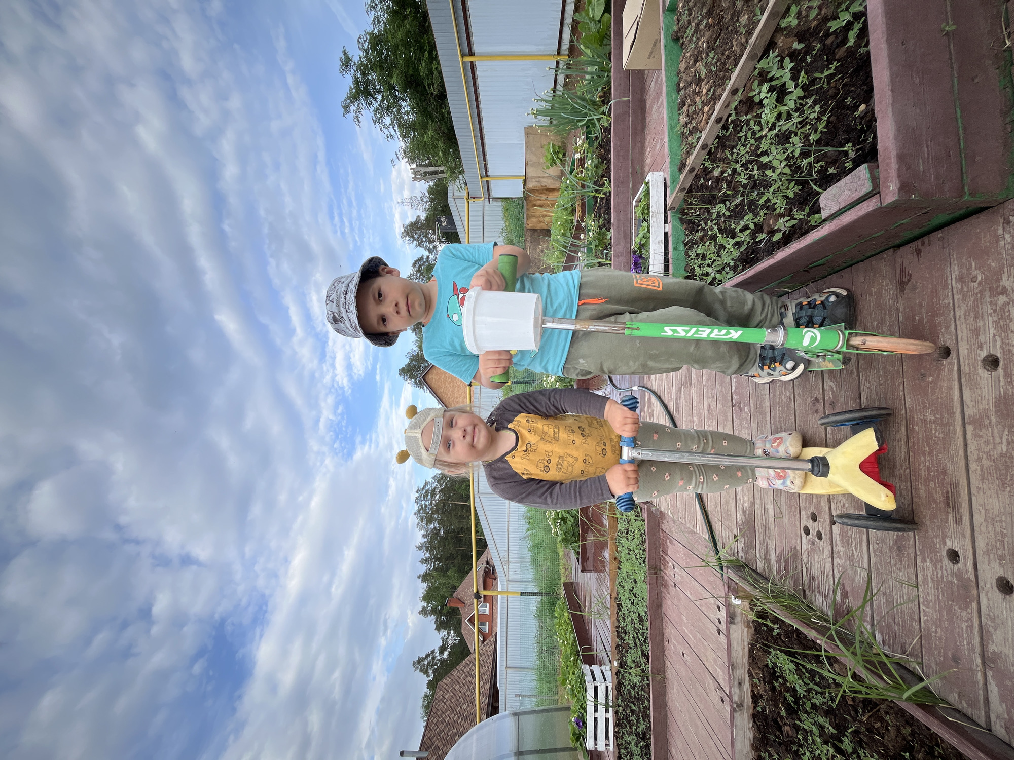 Two children stand on a wooden deck next to raised garden beds. The older child wears a patterned bucket hat, a blue t-shirt, and green pants, and holds a white plastic tube. He is riding a green scooter. The younger child, with a cap decorated with antennae, wears a yellow and gray long-sleeved shirt and is riding a yellow scooter. The backdrop features a blue sky with white clouds, a fence, vegetation, and glimpses of houses, indicating the location is Chaikovsky, Russia.