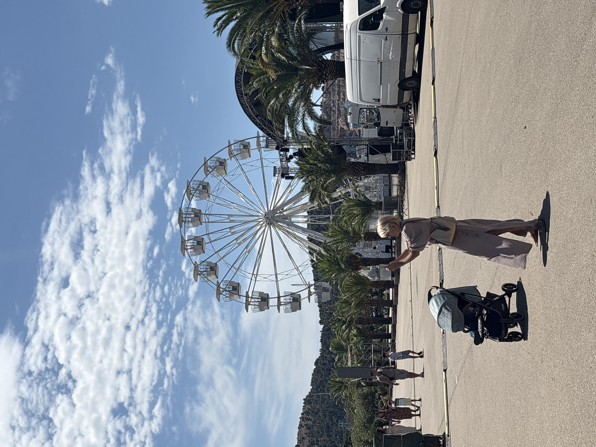 The image depicts an outdoor scene with a large Ferris wheel prominently featured in the background. A woman in a long, light-colored dress is standing in the foreground, facing away from the camera and holding up a smartphone, appearing to take a picture or video. To her left and slightly in front, a baby stroller with a child inside is parked on the asphalt. Several other people, including a family with children, are walking in the middle ground. Palm trees and a stage setup with lighting equipment are visible behind the people. A white van is parked to the right of the frame. The scene appears to be a public area, possibly an amusement park or event venue, with a hilly, vegetated landscape in the far background. The sky is blue with scattered white clouds. The location is identified as the city of Bar, Montenegro.