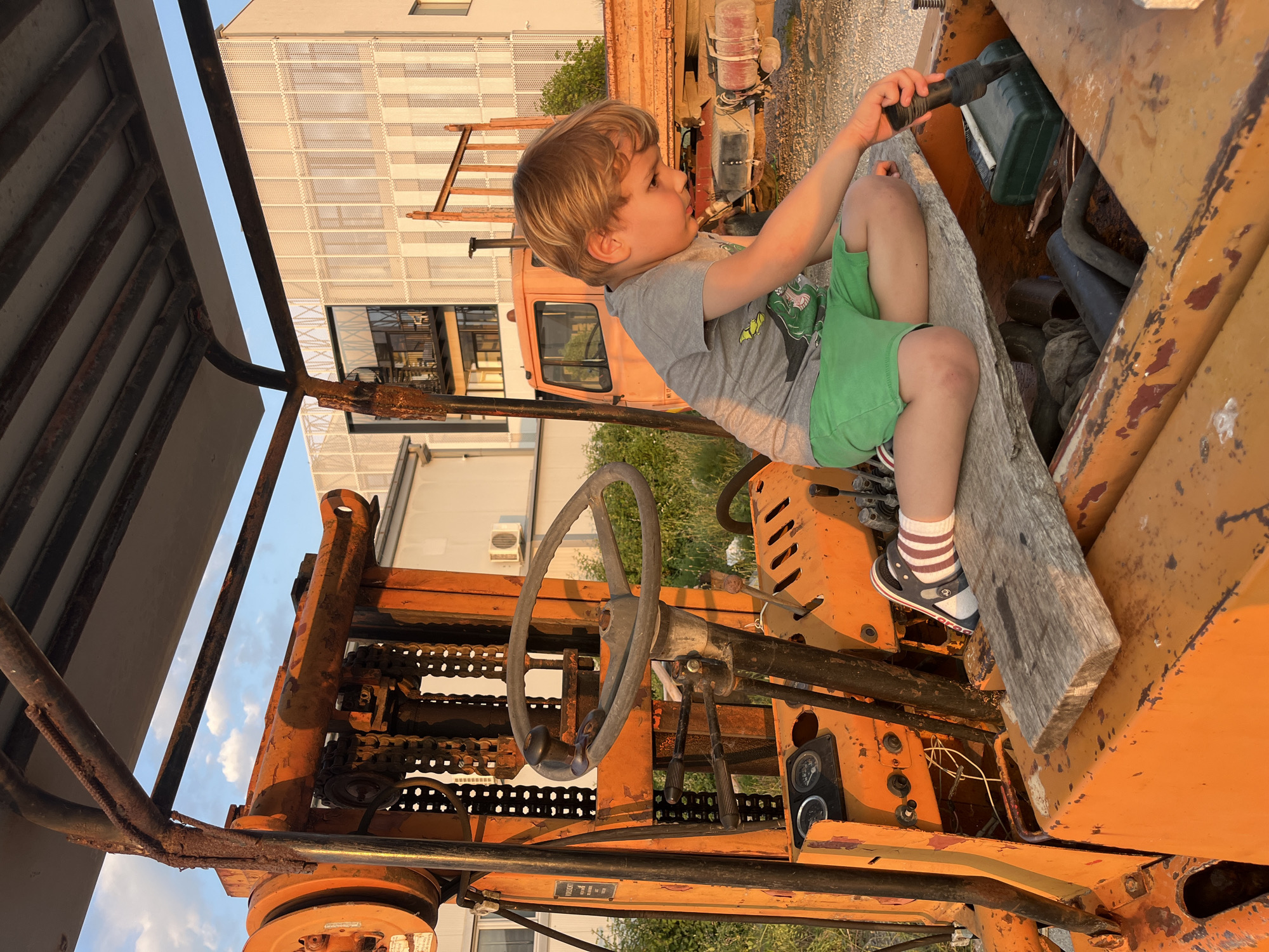 A young boy is sitting on a piece of wood inside the engine compartment of an orange, rusty forklift. He is holding a black object. The background includes a building with large windows and another orange vehicle, potentially a truck. The location is Pobrdje, Montenegro.