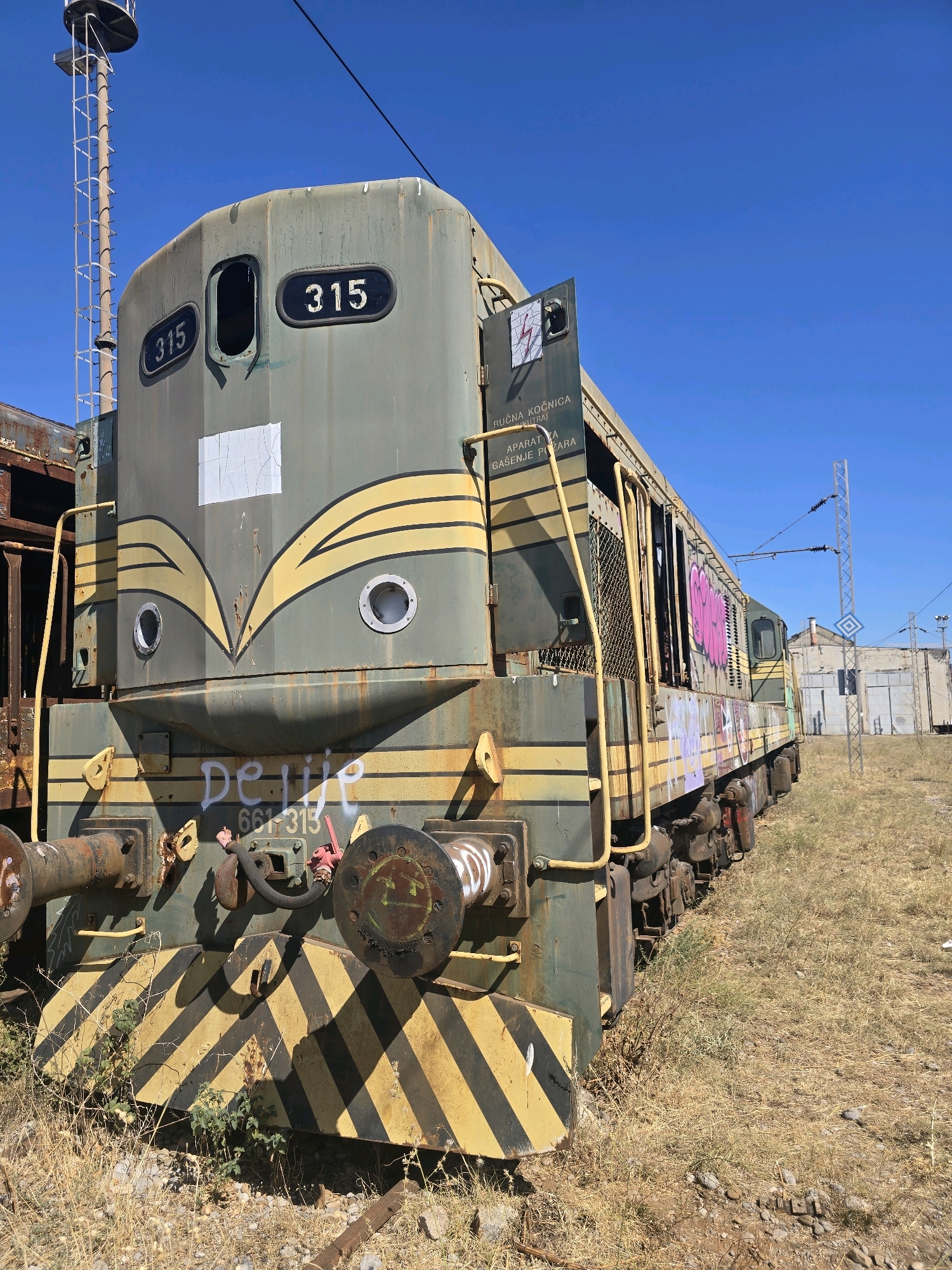 An abandoned, weathered, green and yellow diesel locomotive, identified by the number 315, is parked on a dry, grassy embankment under a clear blue sky. The locomotive shows signs of age and neglect, with rust and peeling paint visible. Graffiti adorns the side of the train. In the background, industrial buildings and a tall electrical pylon are visible. The overall scene suggests a disused railway yard.