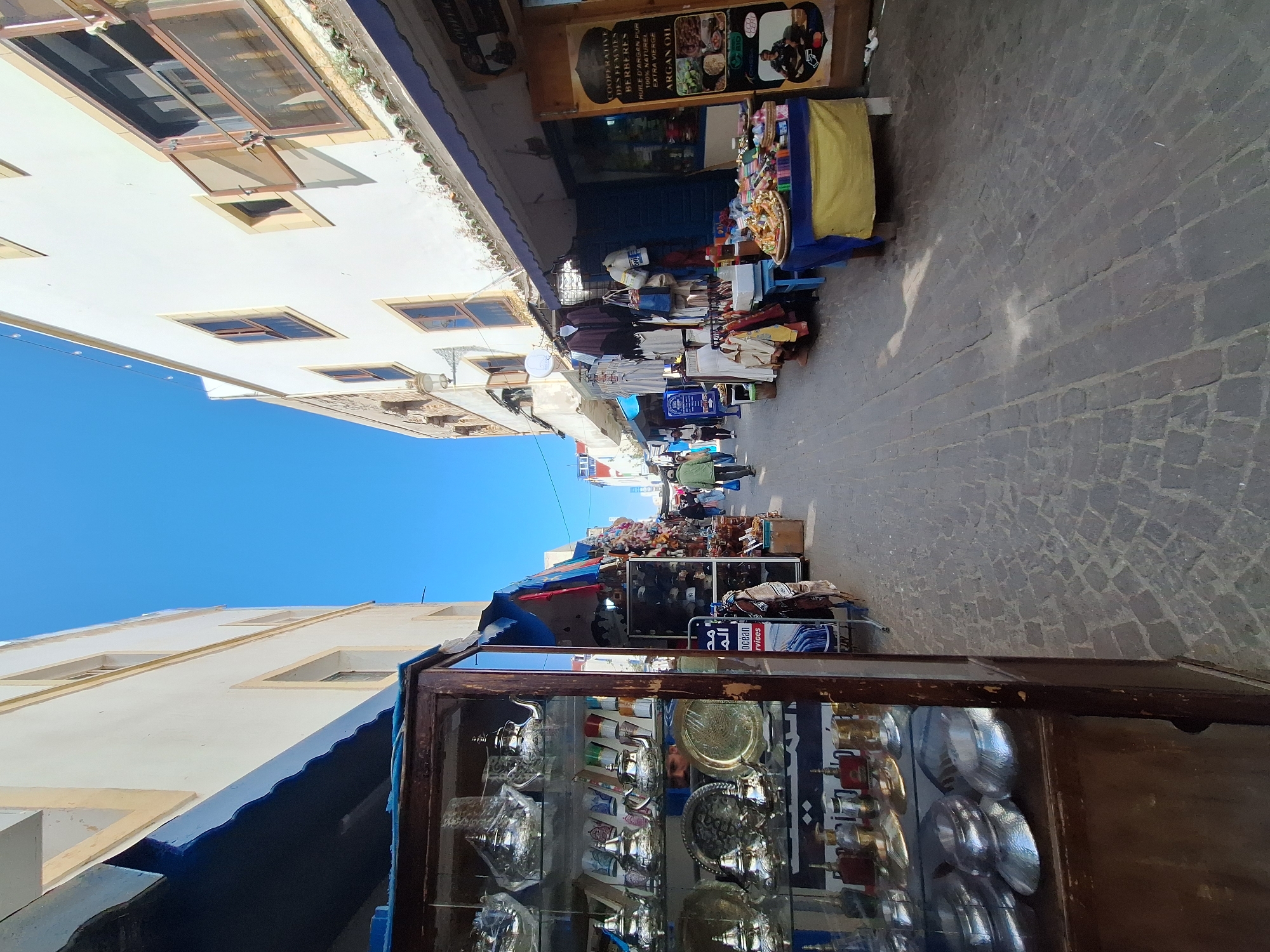 The image captures a narrow street in the Moroccan city of Essaouira. The perspective is taken from a low angle, looking upward towards the sky. Buildings line both sides of the street, with white facades and blue accents.
Stalls and shops are set up along the street, displaying various goods such as silver teapots and plates in glass display cases, clothing, and other souvenirs. People are walking through the street, browsing the merchandise and interacting with vendors.
The street is paved with cobblestones, adding to the historic atmosphere of the area. The sky is clear and blue.