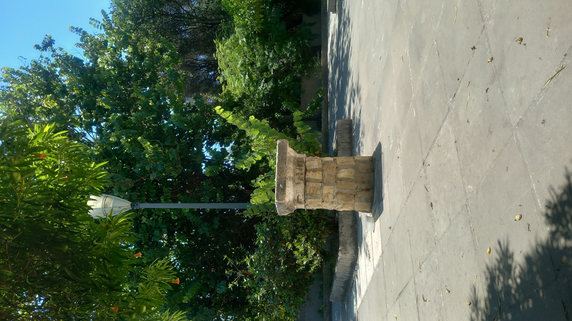 A weathered stone pedestal stands on a paved courtyard in Kos, Greece. The pedestal, constructed from irregular blocks of stone, has a circular base and a flat, octagonal top. A tall, modern lamppost with a white plastic fixture rises behind it. Lush green trees with some orange blossoms surround the scene, casting dappled shadows on the grey, square-tiled pavement. The sky above is a clear, bright blue. The image captures a quiet, sun-drenched corner, likely in a park or public square.