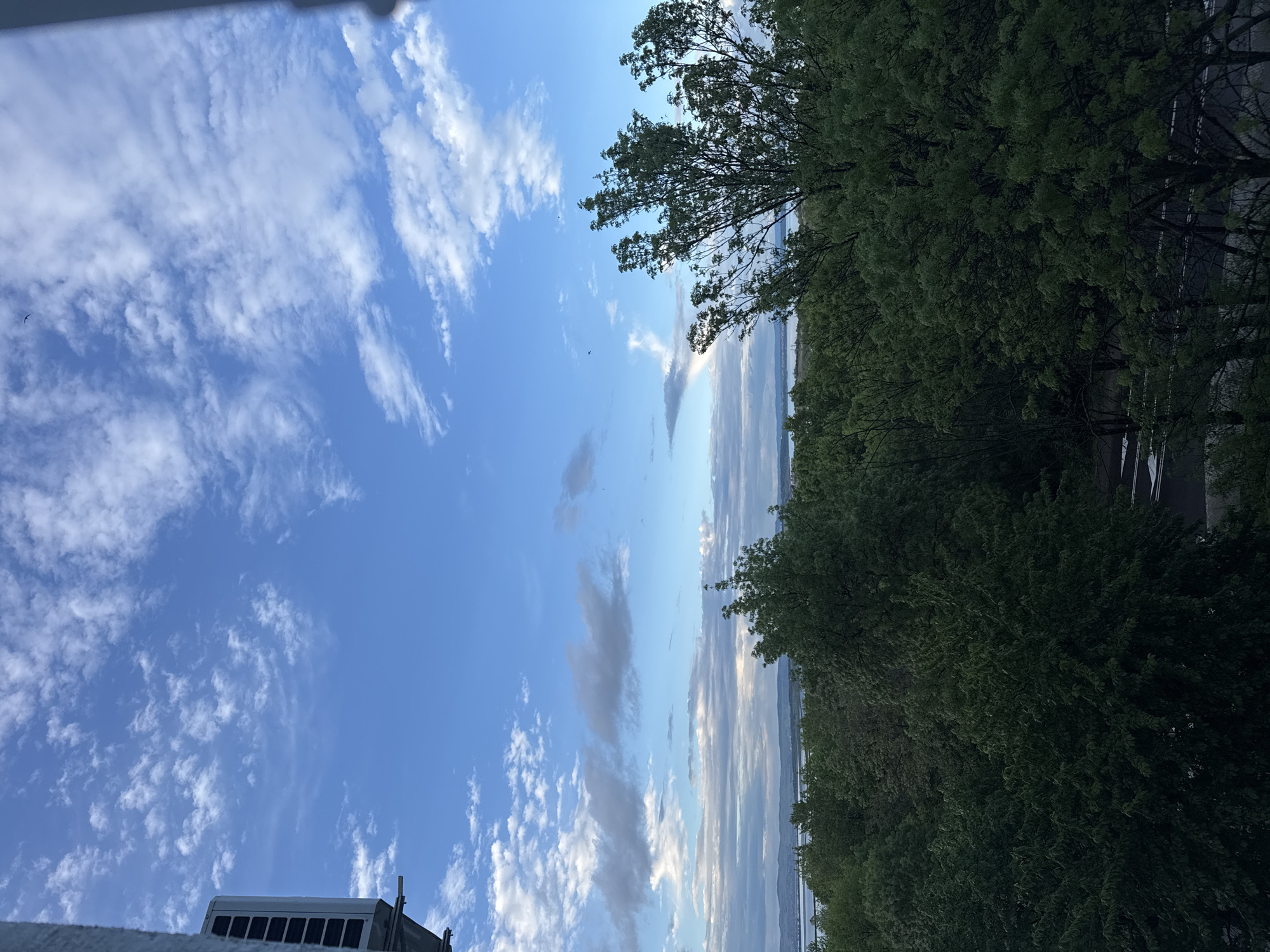 The image shows a view of the sky and a line of trees. The sky is mostly blue with scattered white clouds. There are some long, thin clouds near the horizon. A few trees are visible on the right side of the image. In the bottom left corner, there is a building or structure with some sort of equipment on top. The location is likely near the sea. Due to the type of clouds and trees, it is possible the image was taken in Burgas, Bulgaria, but no specific landmarks are identifiable in the image to confirm the location.