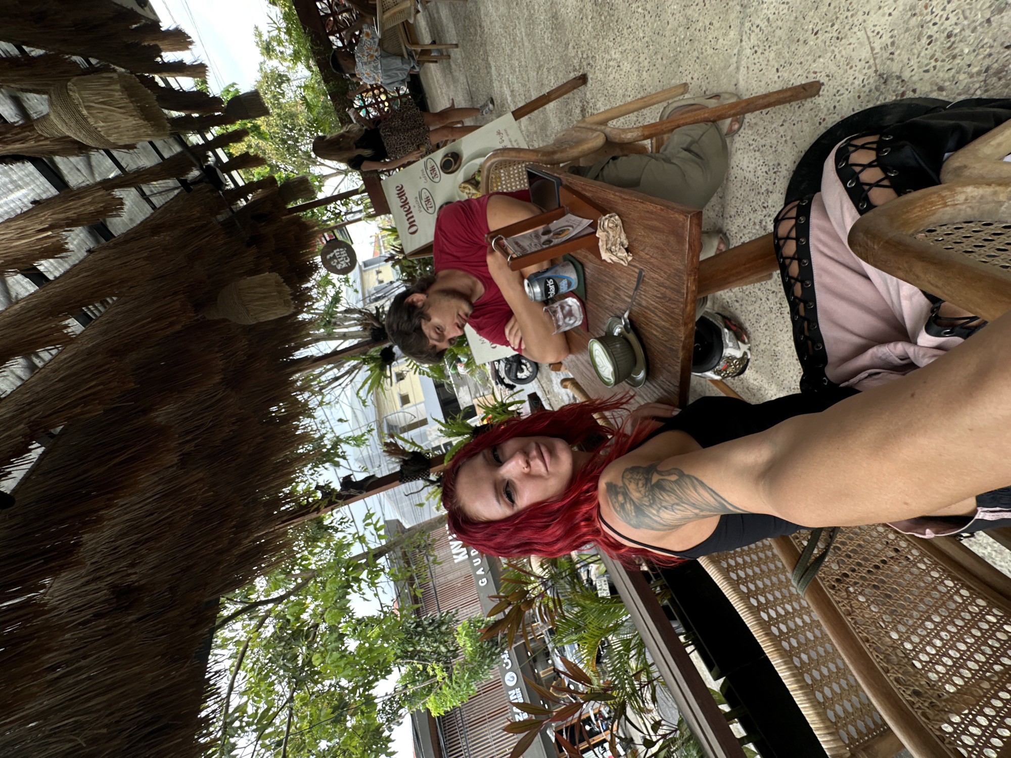 The image depicts an outdoor seating area at a cafe in North Kuta, Indonesia, characterized by a thatched roof and lush greenery. A woman with vibrant red hair, wearing a black tank top and a pink skirt with black lace details, is in the foreground, looking towards the camera. She has a tattoo on her left arm. To her right, at a wooden table, a man in a red t-shirt with his arms crossed appears to be dozing. On the table are a cup of green tea, glasses of water, a can of soda, and a menu board. In the background, another woman in a black top and a leopard print skirt is standing, and a child is seated at a table further back. The cafe appears to be open-air, with wooden furniture and woven accents.