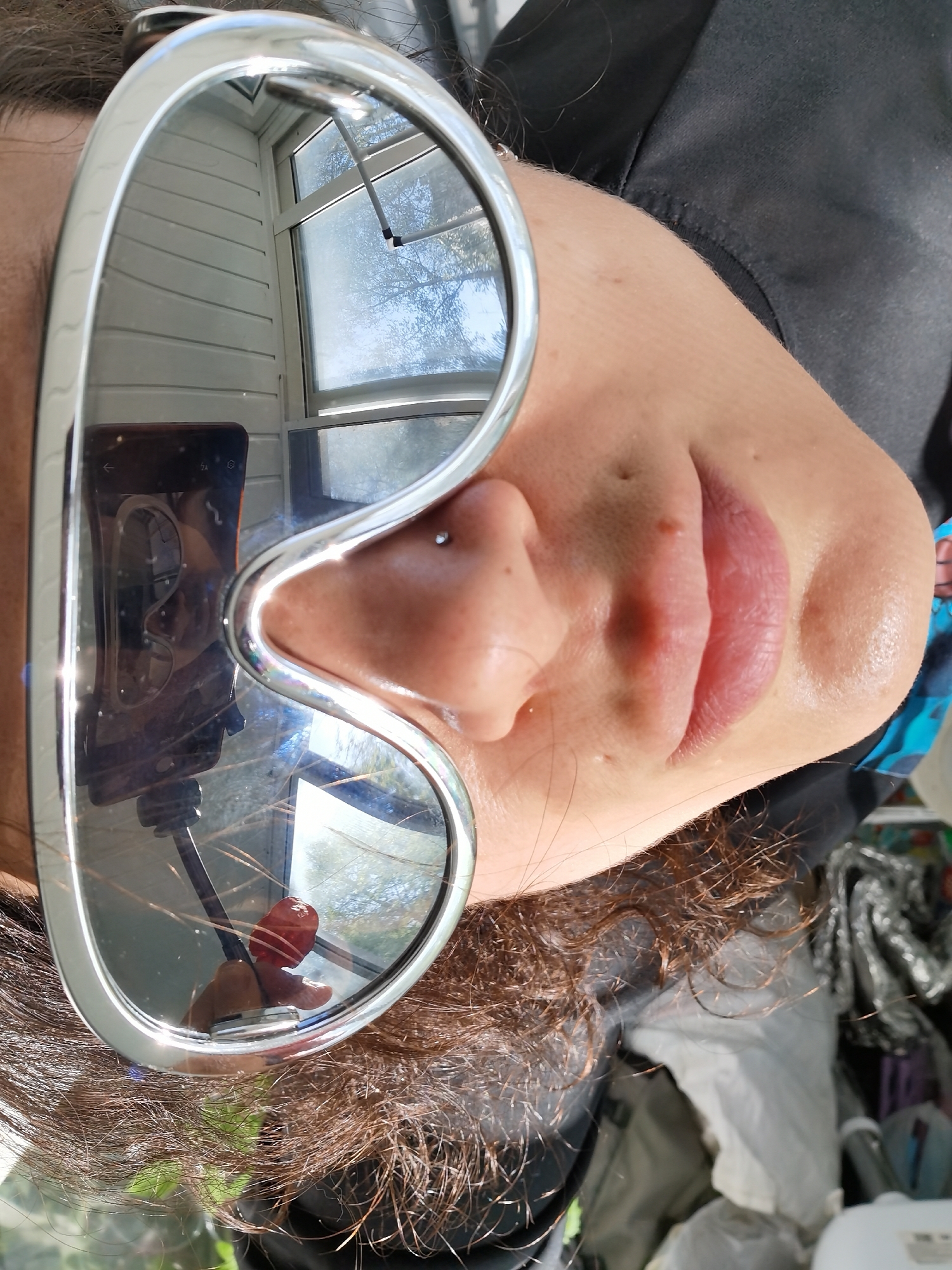 A selfie taken indoors, possibly in Chaikovsky, Russia. A fair-skinned individual with a nose piercing is wearing large, mirrored sunglasses that reflect a window with a view of trees and sky. The reflection also shows a hand holding a red object, possibly a button or a decorative piece, and a phone mounted on a stand. The person's lips are visible, and their curly brown hair is scattered around their head. The background is cluttered with various items, including fabric and containers.
